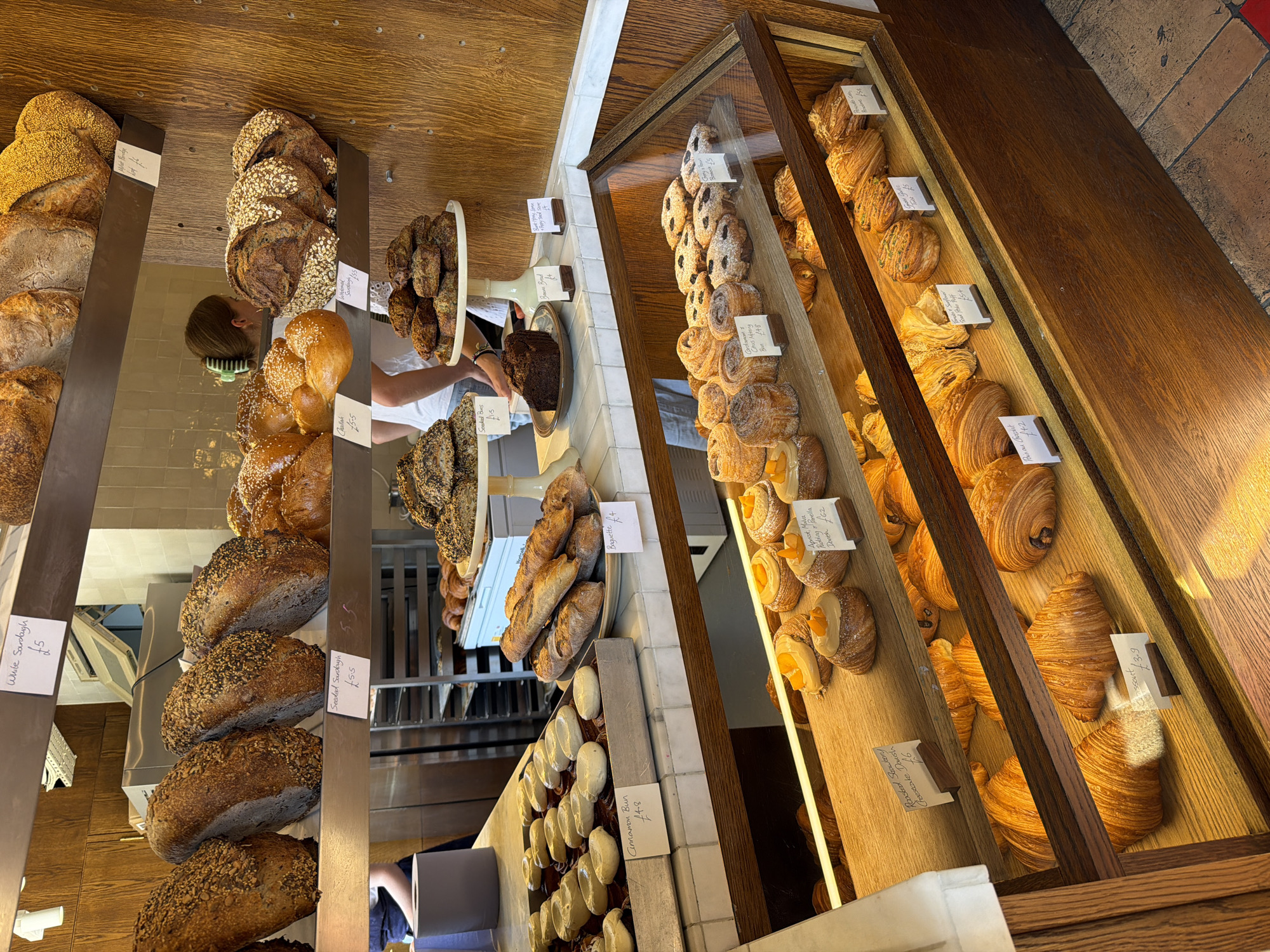 This is an image of a bakery display in London, United Kingdom, showcasing a variety of baked goods. The display shelves are made of wood and marble.
On the top shelf, there are several loaves of sourdough bread, some with seeds, and a few pastry items. Price tags are visible, indicating prices like "£5" and "£5.5".
The middle shelf features more bread, including baguettes, and a dark loaf of banana bread. There are also trays of cinnamon buns and other pastries. Some of these items have price tags indicating "£4", "£4.5", and "£3".
The bottom shelf, enclosed in glass, presents a variety of pastries and tarts. These include croissants, danishes with fruit toppings, and other small, sweet baked items. Price tags visible in this section include "£6", "£5", and "£3.9".
In the background, a person wearing a white shirt and a green hair clip is visible behind the counter, suggesting the bakery is operational. The overall environment is that of a well-stocked bakery, with an emphasis on artisanal bread and pastries.