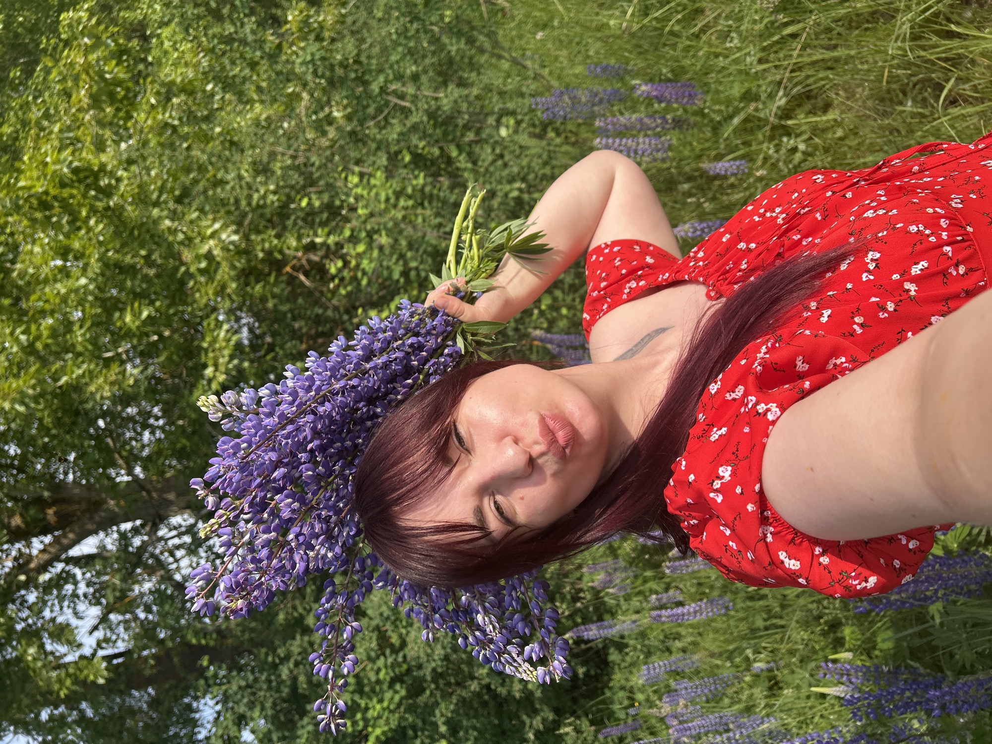 A woman with dark red hair and bangs, wearing a red dress with a white floral pattern, is taking a selfie in a field of purple lupine flowers. She is holding a bouquet of lupine flowers above her head and has her lips puckered as if for a kiss. The background consists of lush green trees and more lupine flowers. The image appears to have been taken outdoors in a natural setting, likely in the city of Garevaya, Russia, given the context. The lighting suggests it is daytime, possibly late afternoon due to the warm tones.