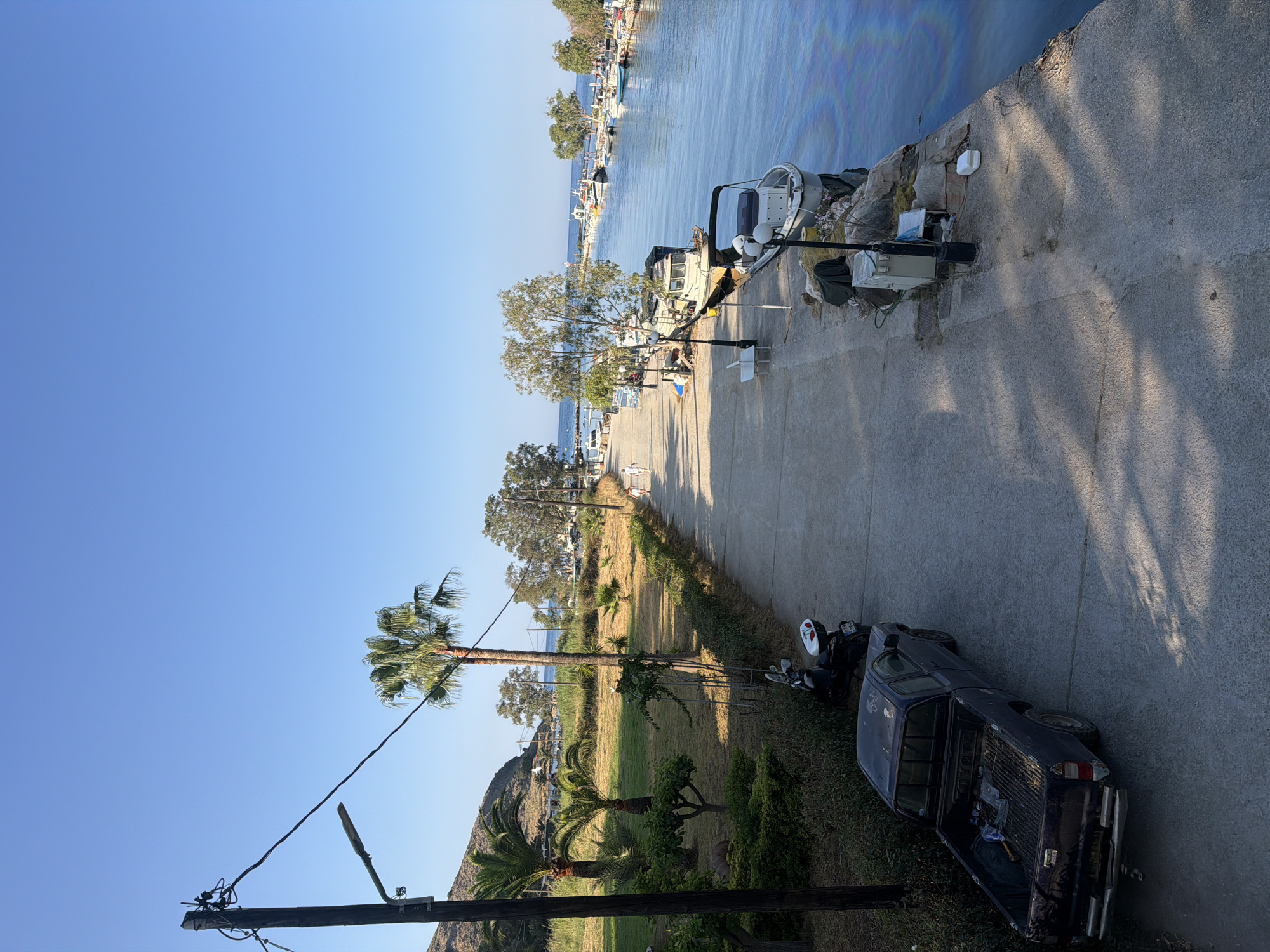 The image depicts a sunny day in Georgioupoli, Greece, showing a paved walkway along a calm body of water. On the left side of the walkway, there is a grassy area with several palm trees and other vegetation, backed by a hill. A dark-colored pickup truck is parked on the walkway, and a scooter is parked behind it. Further down the walkway, there are benches and lampposts. Several boats are docked along the right side of the walkway, in the water. The water exhibits an iridescent sheen, possibly from oil. In the distance, more boats are visible, as well as trees and buildings lining the opposite bank of the waterway. Two individuals are walking on the walkway in the distance. The sky is clear and blue.