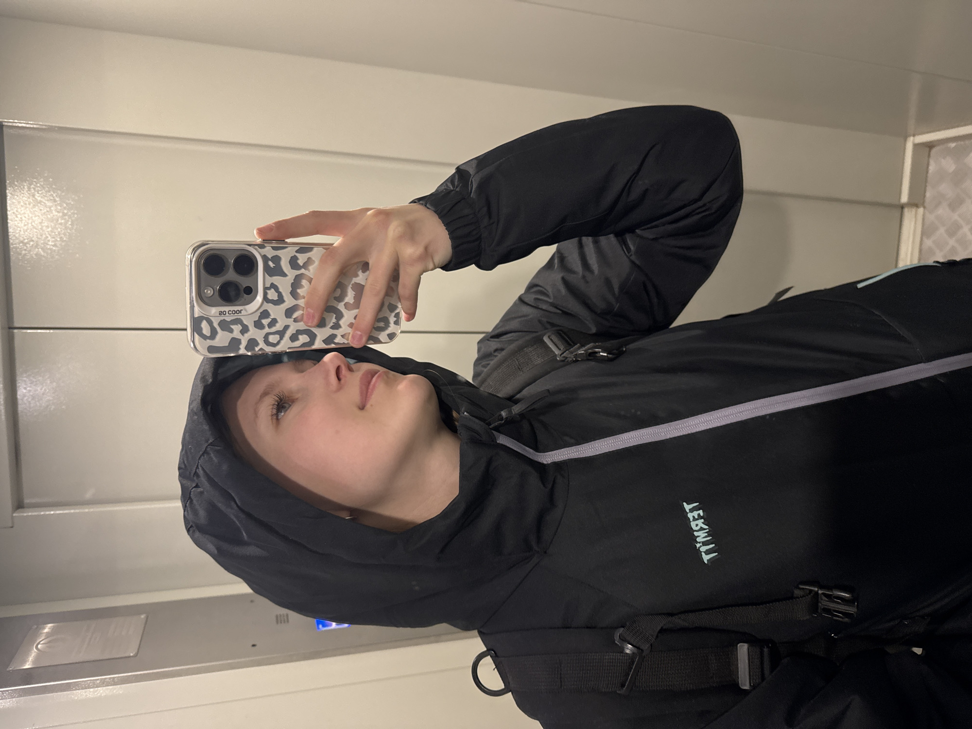 A person is taking a selfie in what appears to be an elevator. The person is wearing a black hooded jacket and has a phone with a leopard print case. The surrounding environment consists of elevator walls. There is no discernible information in the image to indicate its location within Chaikovsky, Russia.