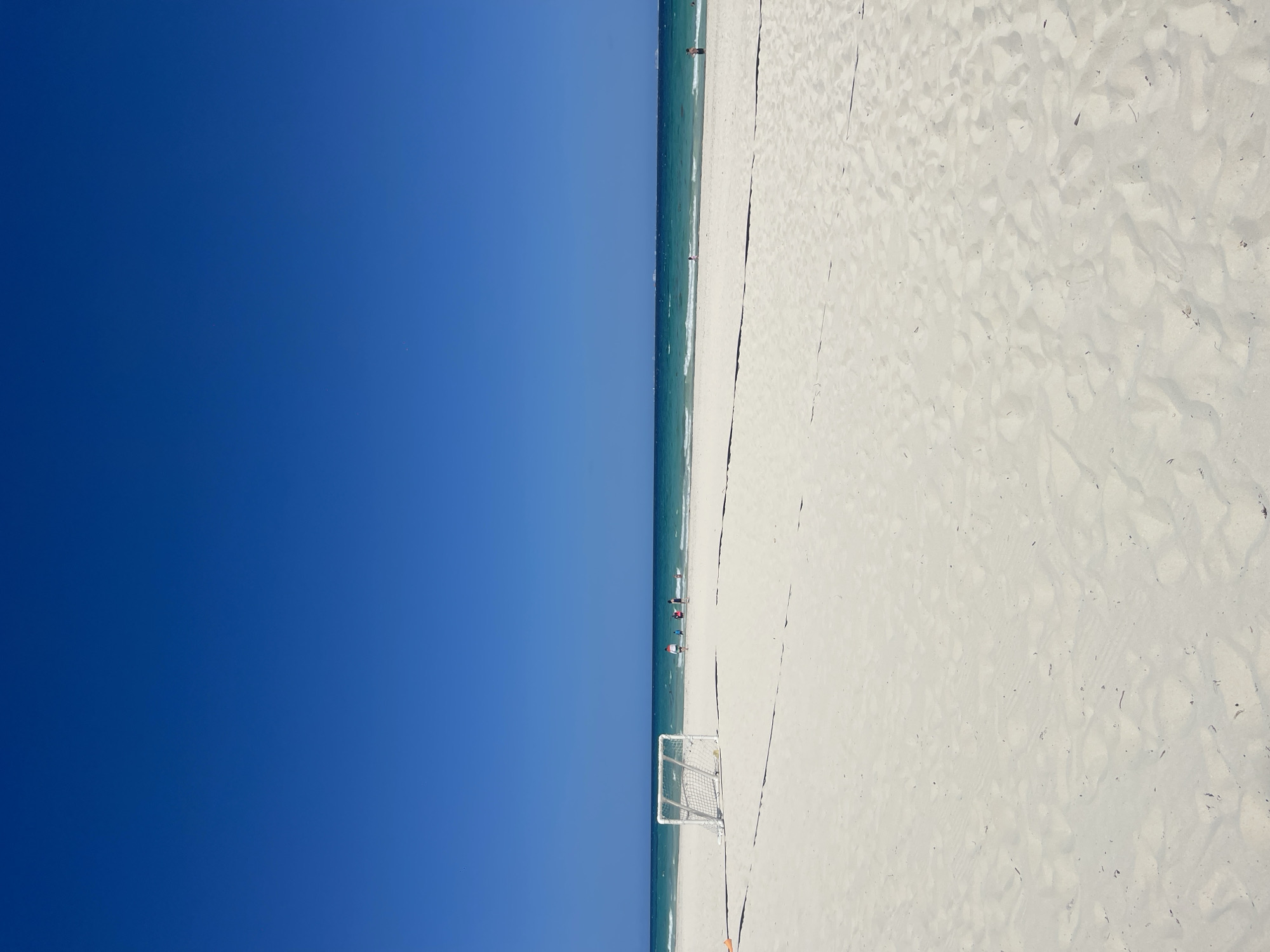 The image shows a beach scene in Playa del Carmen, Mexico. The left side of the image is a clear, blue sky. A narrow strip of turquoise sea divides the sky from the white sand beach on the right side of the image. A small, white soccer goal is visible near the water's edge. Several people are standing in the water and on the shoreline. The sandy beach extends into the foreground.