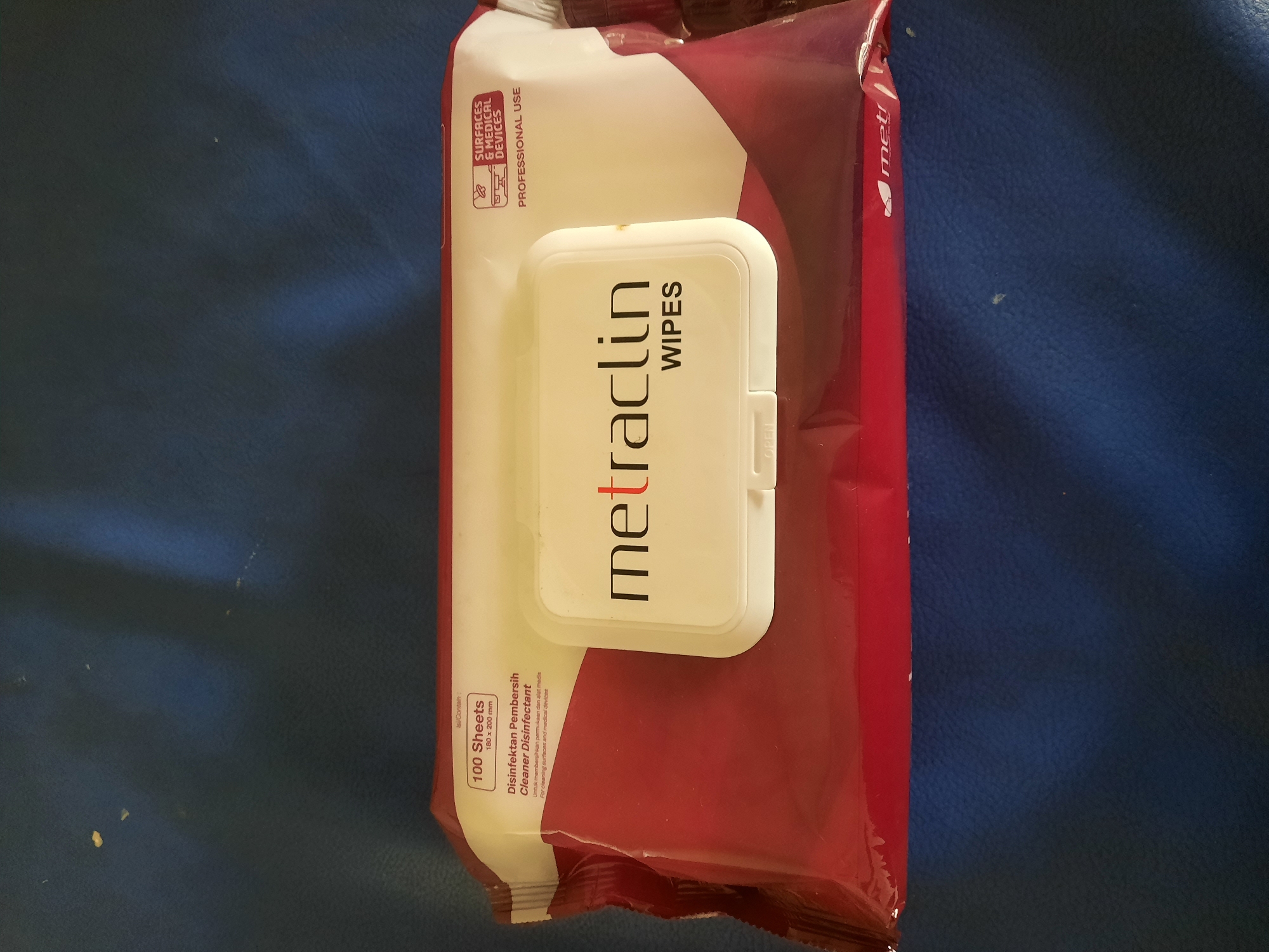 The image shows a package of "metraclin WIPES" disinfectant wipes. The packaging is predominantly red and white, with the brand name "metraclin" prominently displayed in red and black lettering on a white flip-top lid. Text on the packaging indicates "100 Sheets" and "Disinfektan Pembersih Cleaner Disinfectant," along with a subheading "Untuk membersihkan permukaan dan alat medis" (For cleaning surfaces and medical devices). A small icon depicts a computer and medical equipment, with the text "SURFACES & MEDICAL DEVICES PROFESSIONAL USE." The package is placed on a dark blue, textured surface, likely a piece of furniture. There are no people or any discernible elements that would place this image in a specific city like Lhokseumawe, Indonesia, or any other specific geographic location.