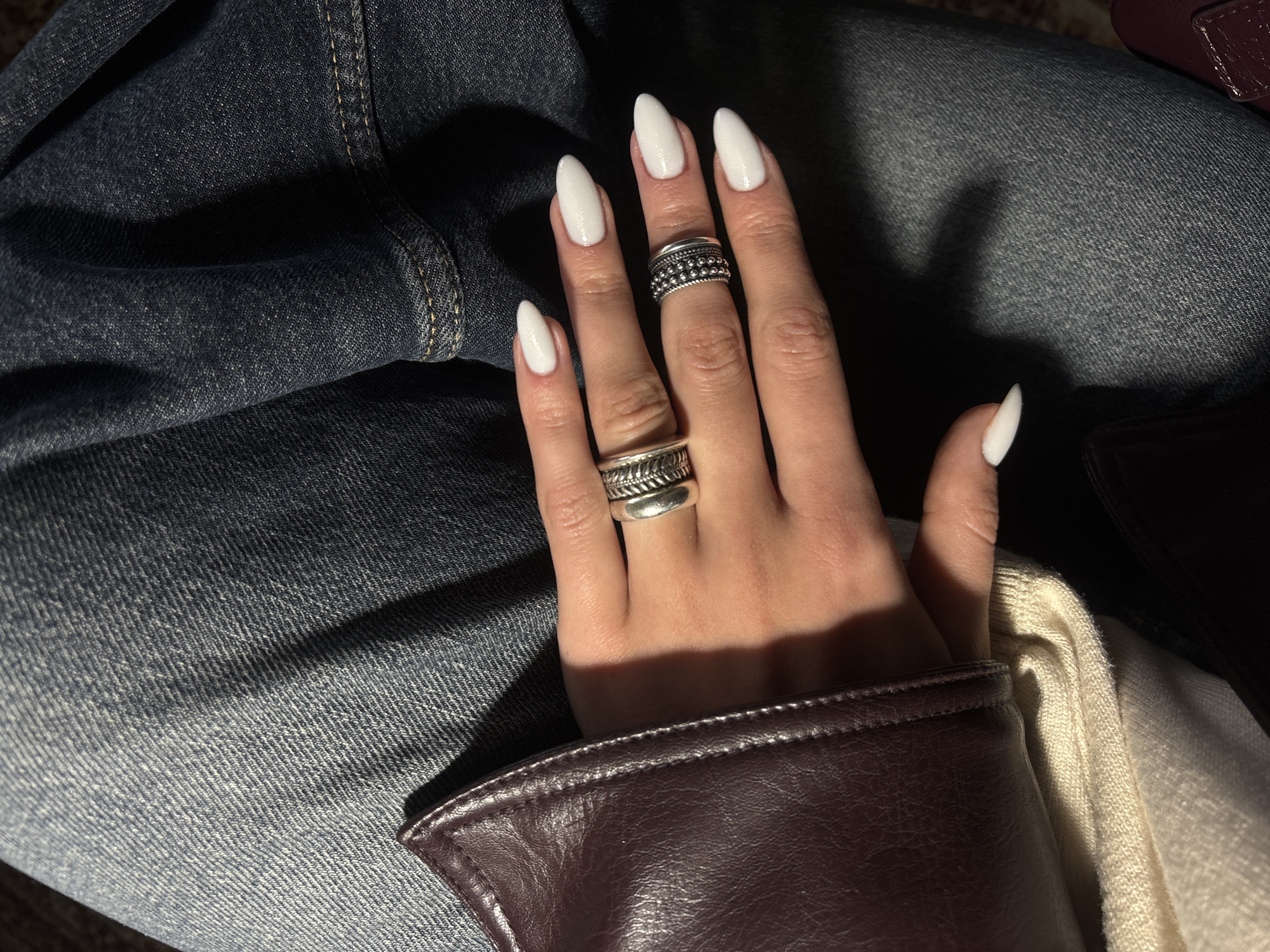 A person's left hand rests on their lap, which is covered in blue denim jeans. The hand has long, almond-shaped white fingernails, and two silver rings are worn, one on the middle finger and one on the index finger. Part of a brown leather jacket or garment is visible near the wrist, with a cream-colored fabric visible underneath it. The location is not discernible from the image.