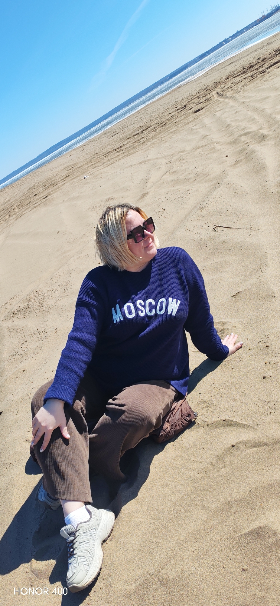 A woman is sitting on a sandy beach under a clear blue sky. She is wearing a dark blue sweater with the word "MOSCOW" in white letters, brown corduroy pants, white socks, and gray sneakers. She is also wearing large, dark sunglasses. The beach appears to be located in the city of Noviy, Russia, based on the sweater. The woman is looking to her right and seems to be enjoying the sunshine. The beach stretches out towards the horizon, where the sky meets the sea. The camera model of the photograph is Honor 400.