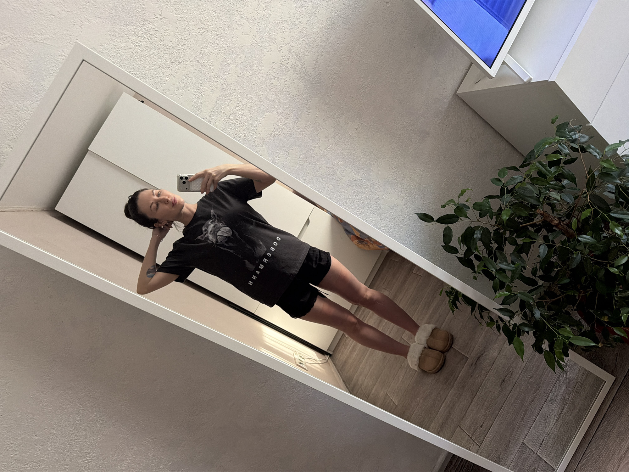A woman is reflected in a full-length mirror in what appears to be an indoor setting. The woman is holding a smartphone, presumably taking a photo of herself. She is wearing a dark t-shirt with some text, black shorts, and tan-colored slippers. In the background, there is a white wall, a white cabinet, and a green plant in a pot. The location is likely in the city of Perm, Russia.