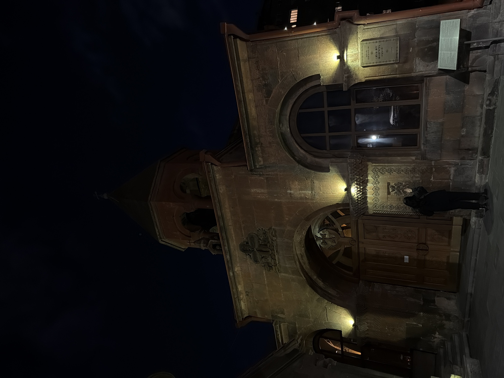 At night, a man stands in front of Saint Anna Church in Yerevan, Armenia. The church's facade is lit by several spotlights. The man is looking toward the church entrance. The sky above is dark. The church is constructed of stone, with rounded windows and archways. A sign is posted next to one of the windows.