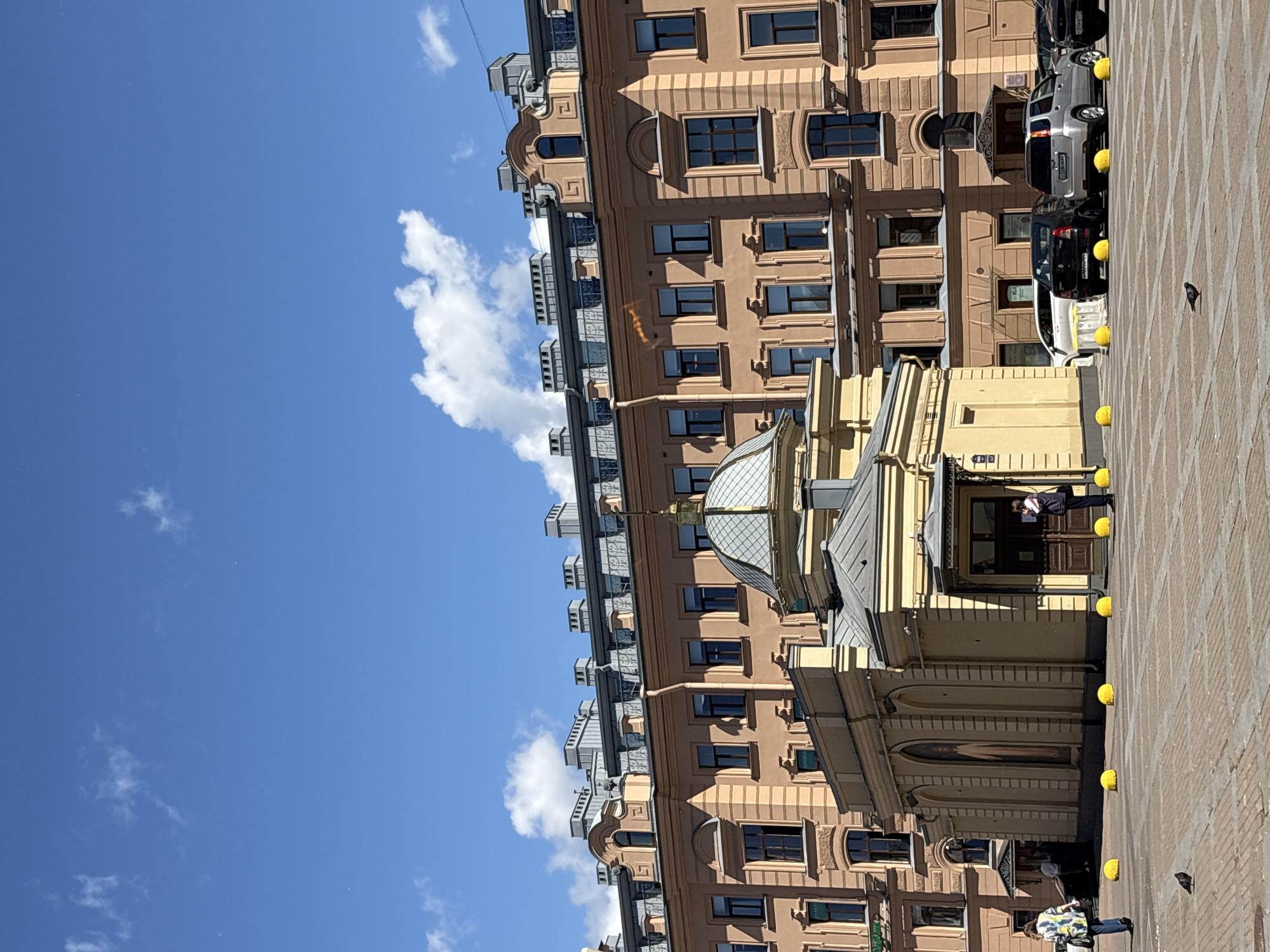 The image shows a bright, sunny day in Saint Petersburg, Russia, with a clear blue sky and scattered white clouds. The scene is dominated by the exterior of a building with a salmon-pink facade, featuring multiple stories of windows and decorative architectural elements. A smaller, yellow structure with a domed roof is situated in front of the larger building.
In the foreground, there is a paved square with yellow spherical bollards placed at intervals. Several cars are parked in the background, suggesting an urban setting. Two individuals are visible: one person walking away from the camera on the left, and another person standing near the entrance of the yellow structure on the right. A pigeon is also present, standing on the paved ground. The overall impression is of a public space in a historic city.