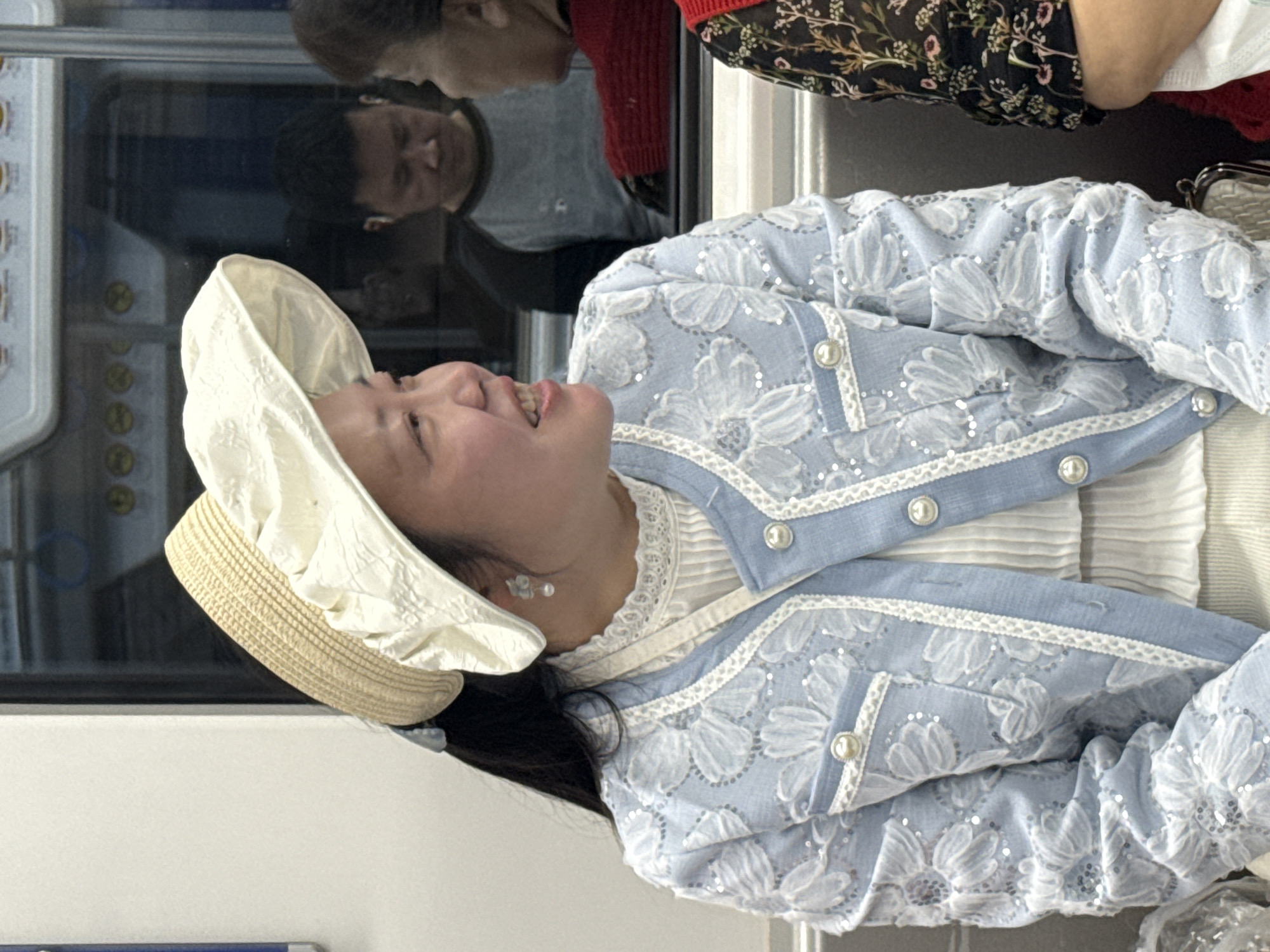 A woman wearing a light blue floral jacket, white dress, and a wide-brimmed straw hat with white fabric embellishments is seated in what appears to be a subway car. The woman is looking up and smiling. The reflection in the subway car window shows two other people; one individual is wearing a grey shirt and the other is wearing a black floral shirt with a red top. The subway car interior is visible through the window, featuring a control panel and other details. The location is likely within Jiulongpo District, China based on the user's query.
