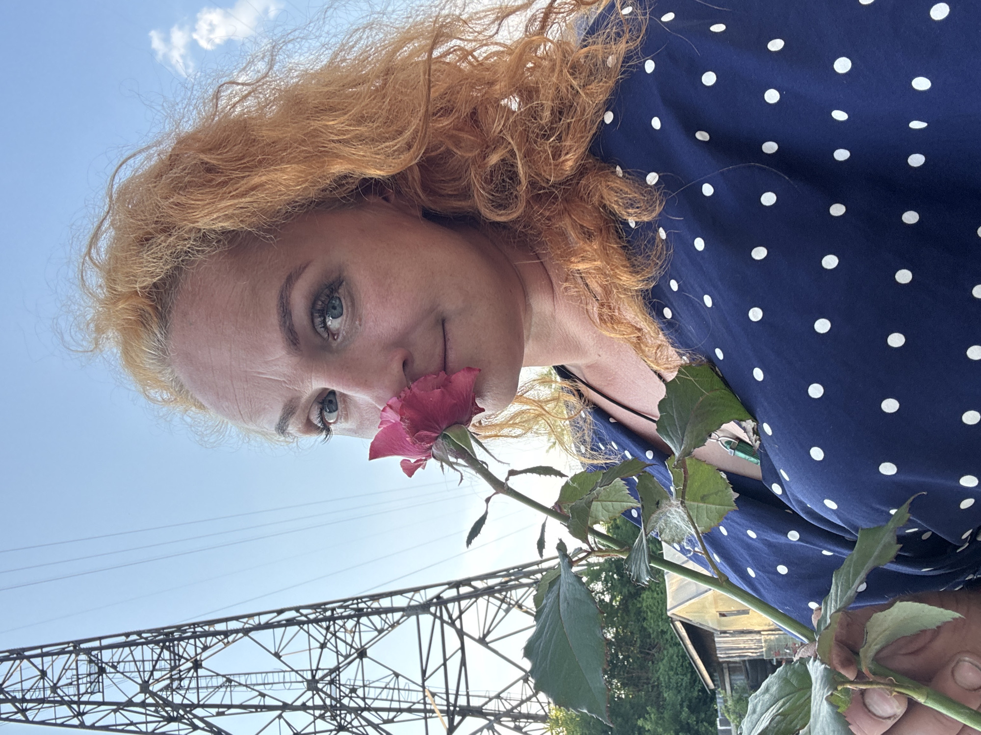 In Chaikovsky, Russia, a woman with curly red hair and blue eyes is captured in a close-up portrait, holding a pink rose and smelling it. She wears a dark blue dress adorned with white polka dots. Behind her, a tall, industrial metal tower with power lines dominates the background, silhouetted against a clear blue sky with a few wispy clouds. The environment appears to be outdoors, possibly in a semi-rural or industrial area of the city, with hints of vegetation and small structures visible in the distance. The woman's gaze is directed towards the camera, and she has a subtle smile. The image is taken from a low angle, emphasizing the imposing structure behind her.
