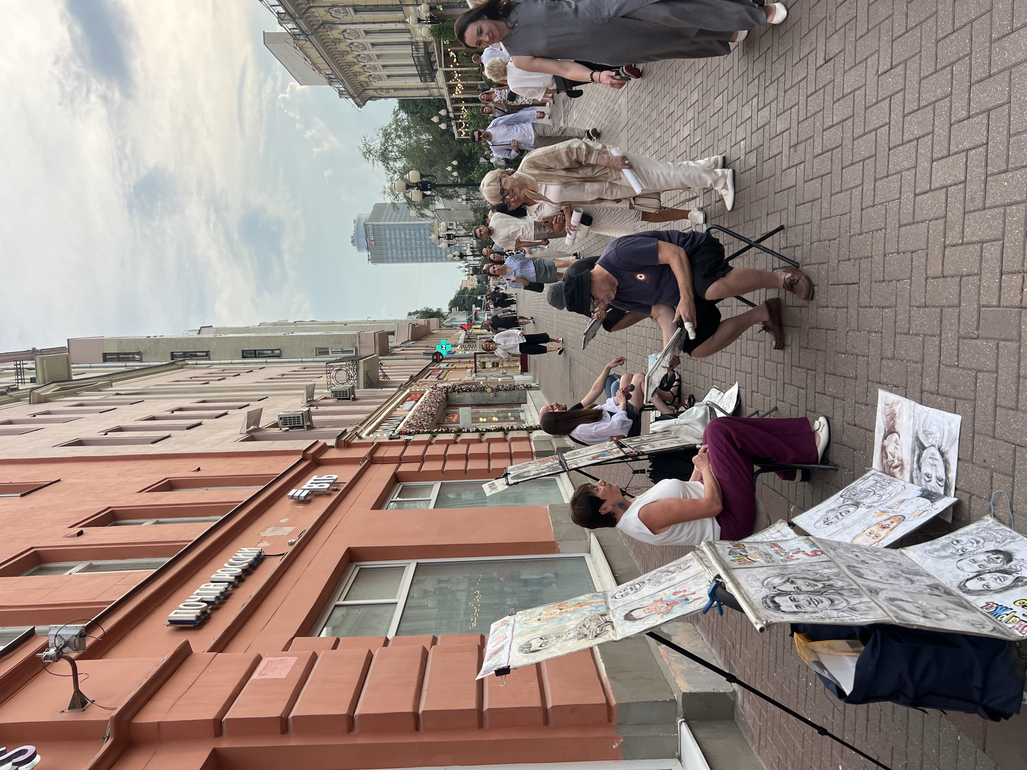 The image depicts an outdoor street scene in Moscow, Russia, featuring several people engaged in drawing. The perspective is from above, looking down onto a brick-paved sidewalk alongside a building. The overall scene is outdoors, likely a pedestrian area or street market.
The subjects are primarily people, some actively drawing and others observing or walking by. There are at least five individuals who appear to be artists, seated or standing with easels and drawing materials. One person is sketching a portrait, holding a pencil and a drawing pad. Several completed drawings, mostly portraits of people, are displayed on stands and on the ground.
The activity taking place is the creation and display of art, specifically portrait sketching. The artists are working on their pieces, and their finished work is presented for potential buyers or viewers. The presence of easels, drawing pads, and displayed artwork indicates an artistic event or a regular setup for street artists.
Notable details include the brick pavement, typical of many urban areas. The buildings lining the street suggest a commercial or residential area, with storefronts and windows visible. The lighting suggests it is daytime, possibly late afternoon given the shadows, and the sky is partly cloudy, indicating fair weather. There is a sign visible on one of the buildings, but it is partially obscured and difficult to read clearly due to the angle and resolution, though it appears to be in Cyrillic script. The overall mood appears relaxed and creative, with people engaged in artistic pursuits on a pleasant day.