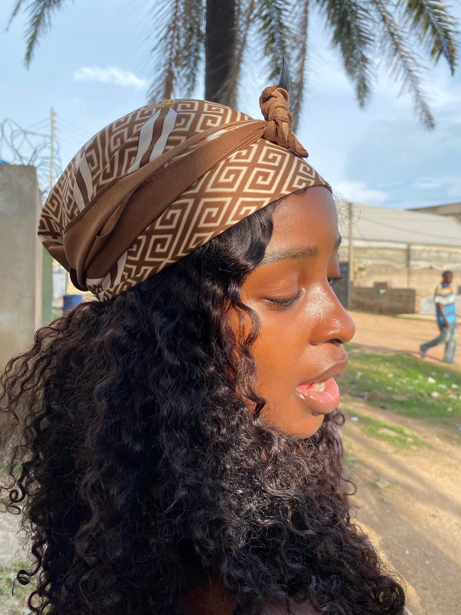 A young Black woman, with dark, curly hair and a brown and white patterned headscarf tied at the top, is pictured in profile against an outdoor background in Sabon Tasha, Nigeria. Her eyes are closed, and her mouth is slightly open, with her lips appearing glossy. Sunlight highlights her face, particularly her nose and cheeks. In the blurred background, a palm tree is visible, along with barbed wire fence, a building, and a person walking. The ground is covered in dirt and scattered debris. The scene appears to be a casual outdoor setting.