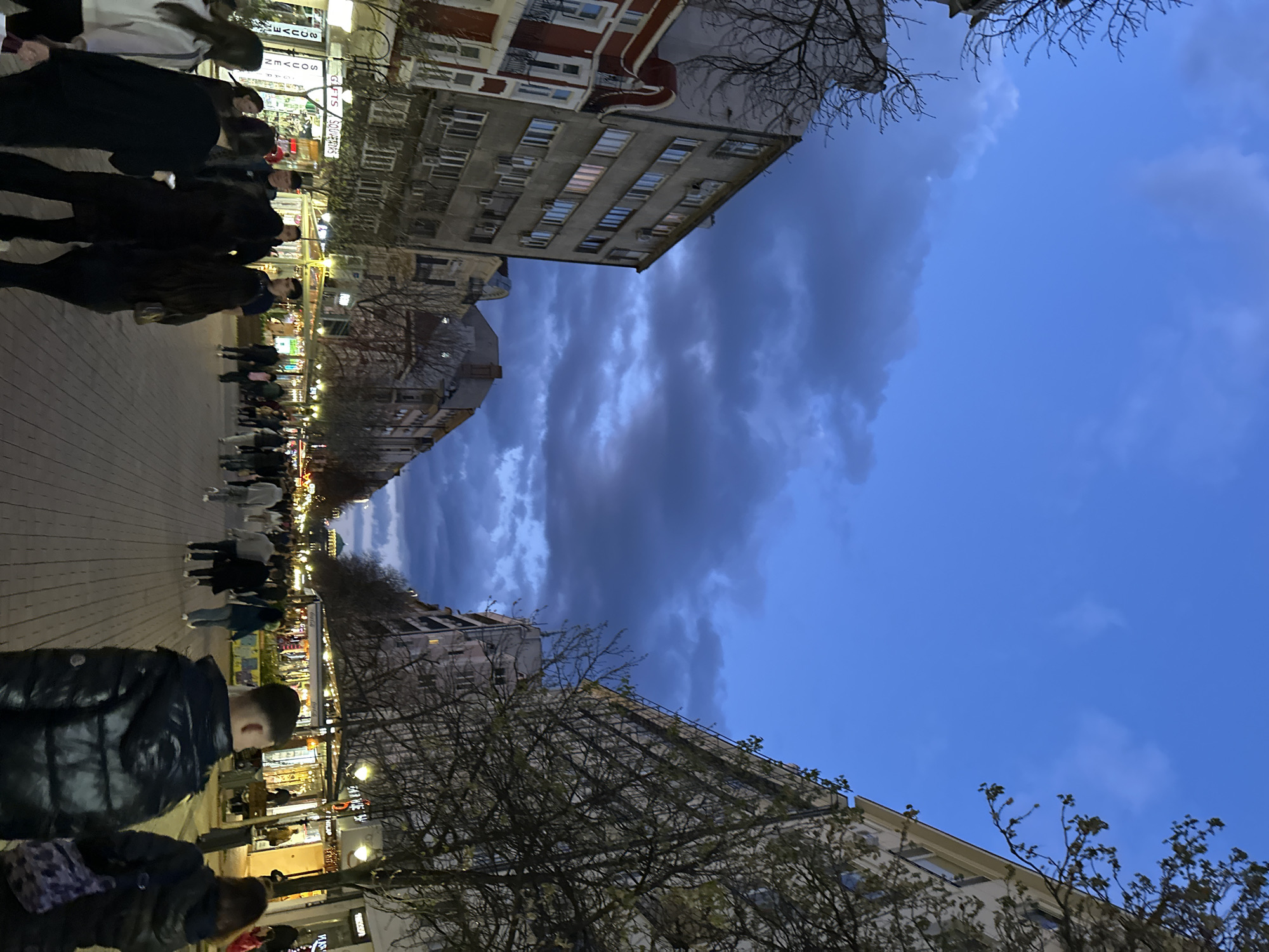 The image depicts a street scene in Sofia, Bulgaria, at dusk. The sky is predominantly blue with patches of dark clouds. Buildings line both sides of the street, and bare tree branches are visible. People are walking along a paved walkway. Shops with illuminated signs are present on the ground floors of the buildings.