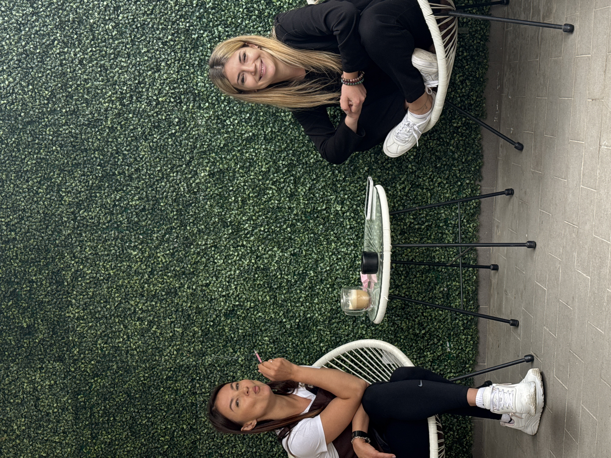 Two women sit at a small outdoor cafe table against a green wall possibly covered in artificial foliage. The woman on the top is blonde and wears black clothing and white sneakers. She is smiling. The woman at the bottom has dark hair and wears a white t-shirt, a brown vest, black leggings, and white sneakers. She holds a pink object, perhaps a straw or pen, near her mouth. The table between them holds a drink, possibly a coffee, in a glass, and a small, dark object, maybe a candle or condiment container. The scene suggests an urban setting, potentially Budva, Montenegro, given the request for the city.