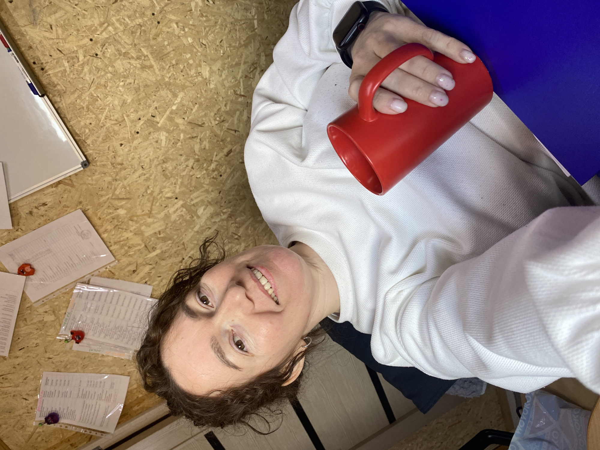 The image displays a close-up, high-angle selfie of a smiling adult female in an indoor setting, identified by the prompt as Ol'khovskoye, Russia; however, no specific visual identifiers within the image corroborate this geographical location. The subject, positioned centrally, has fair skin, brown curly hair, and brown eyes, looking directly at the camera with a wide smile revealing her teeth. She wears a white long-sleeved sweatshirt and small silver hoop earrings. Her right hand holds a red ceramic mug, with her fingernails showing light pink polish, some featuring small red dots. A black smartwatch with a black band is visible on her left wrist.
The background consists primarily of a wall paneled with oriented strand board (OSB). Several documents, some protected by clear plastic sleeves, are pinned to this wall, secured by various colorful, decorative pins, including two prominent ones resembling small red and purple creatures or fruits. A portion of a white magnetic whiteboard with a silver frame and visible markers is present in the upper right. To the left, part of a dark-colored door with two horizontal light-colored stripes is visible. A dark blue folder or notebook rests on a surface in the lower right foreground.