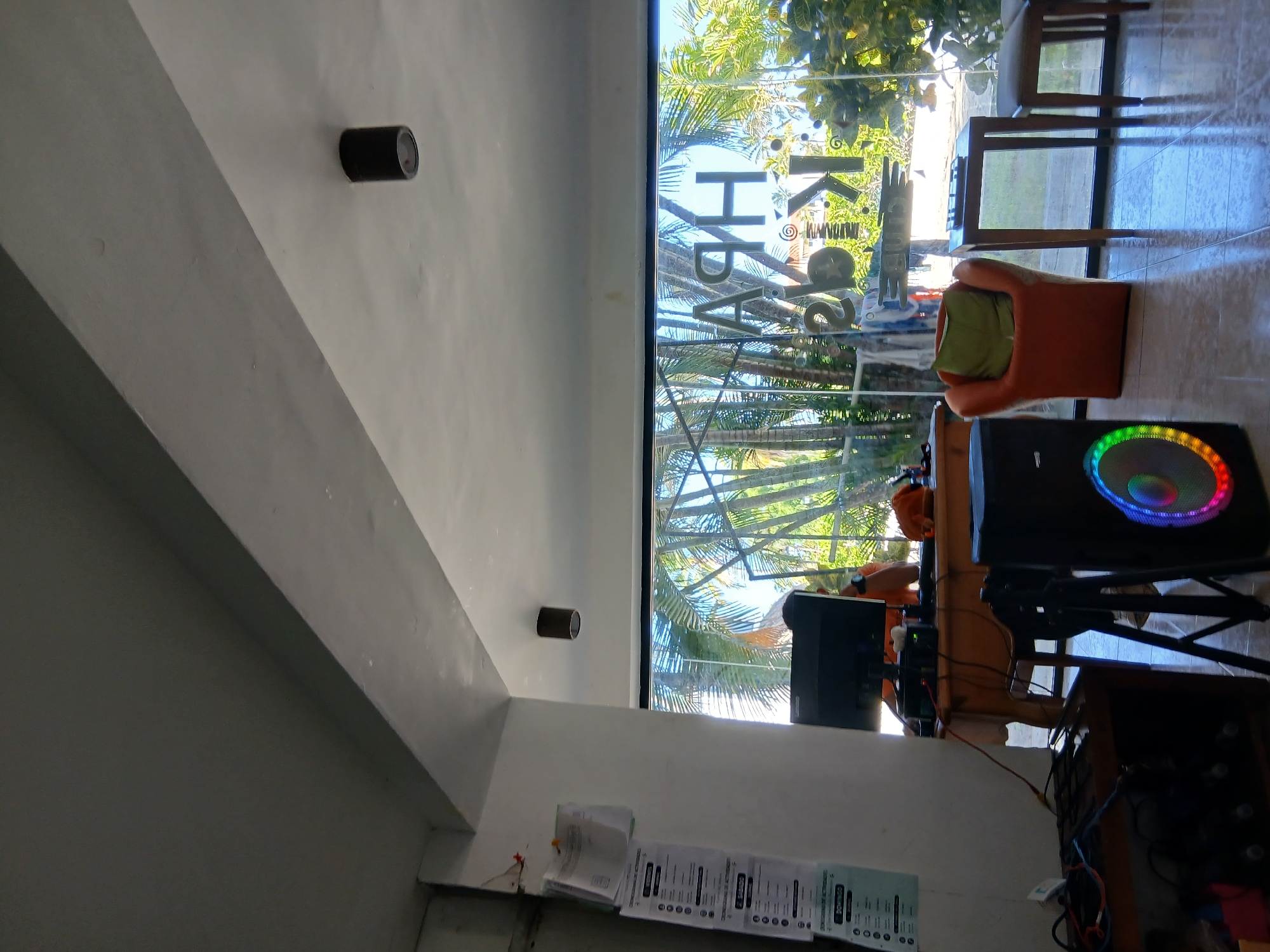 The image depicts the interior of a reception area or office space, likely part of a business located in Playa El Agua, Venezuela. The scene is brightly lit, with natural light streaming in through a large glass window at the back, which offers a view of lush palm trees and greenery.
In the foreground, a tall, black speaker with a circular, multi-colored light display is positioned. To the left of the speaker, a tripod is visible. Further left, there's a dark wooden cabinet filled with audio equipment, including several rows of what appear to be amplifiers or rack units. Cables are neatly organized on top of this cabinet.
A wooden desk occupies the middle ground, on which a computer monitor, keyboard, and other electronic devices are placed. A person, partially visible, is seated behind the desk, wearing an orange shirt.
An orange armchair with a green cushion is situated to the right of the desk, in front of the large window. Adjacent to the armchair is a small, wooden side table with a remote control on it. To the right of the side table, a wooden stool or bench is partially visible.
On the left wall, a bulletin board or wall display with several documents and charts is mounted. The walls are painted white, and the ceiling features recessed lighting fixtures. The floor appears to be tiled and reflective, indicating a polished surface.
The signage on the glass window, though partially obscured and reversed, appears to read "HPA Kids Club" or a similar designation, suggesting the establishment's purpose. The overall impression is that of a welcoming and functional space, possibly for entertainment or services catering to children, situated in a tropical environment.