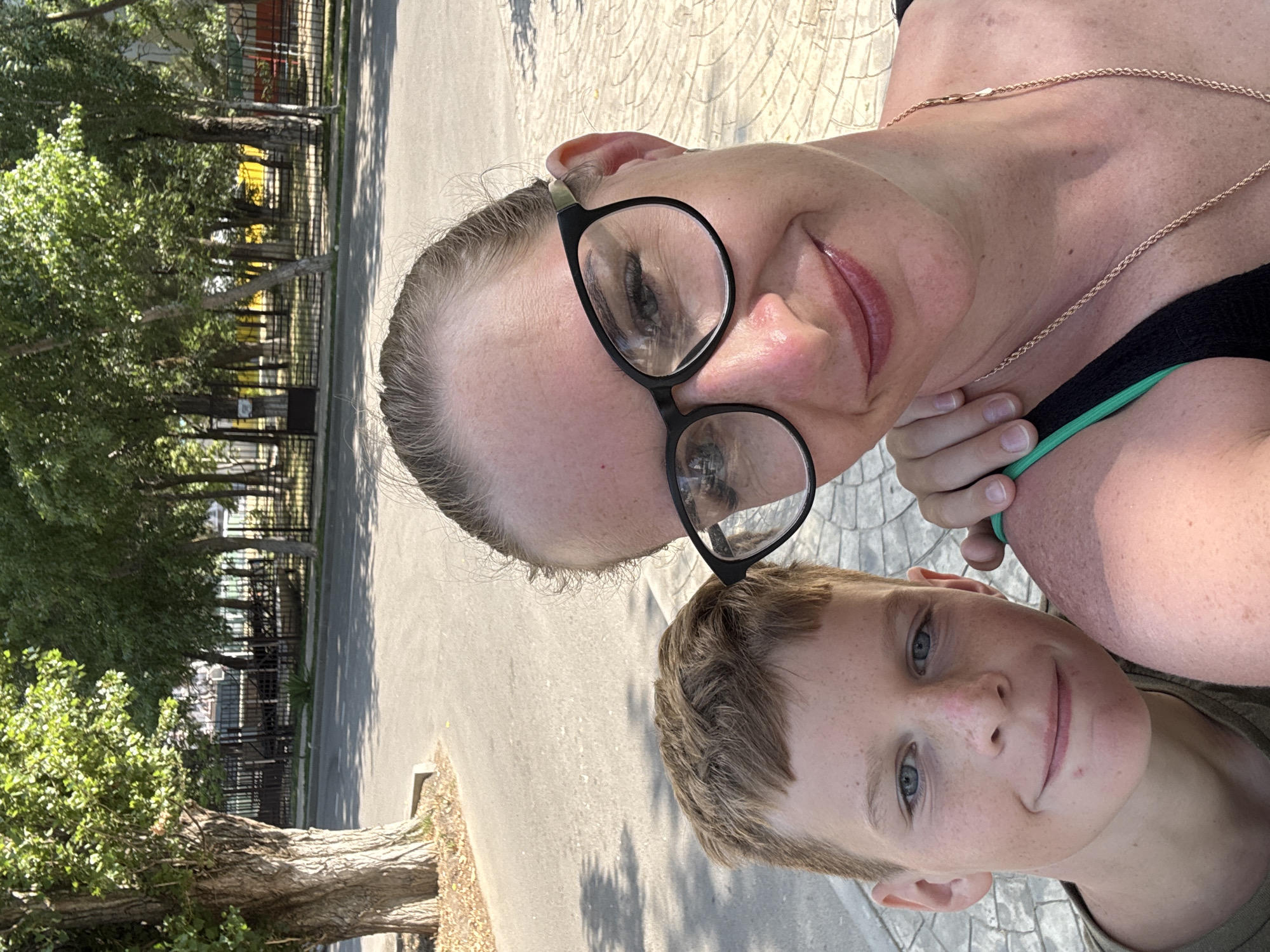 A close-up selfie shows a young boy and a woman, likely mother and son, standing outdoors on a sunny day. The boy, in the foreground to the left, has light brown hair, blue eyes, and freckles. He is wearing a green t-shirt. The woman, on the right, is wearing glasses and a black top with a green strap visible. Her hand rests on the boy's shoulder. Behind them, a paved area with a textured pattern is visible, and beyond that, a street lined with trees and a fence. The background suggests a park or a residential area in a city. The overall lighting indicates it's daytime, with shadows cast on the ground. The location is identified as Vityazevo, Russia.