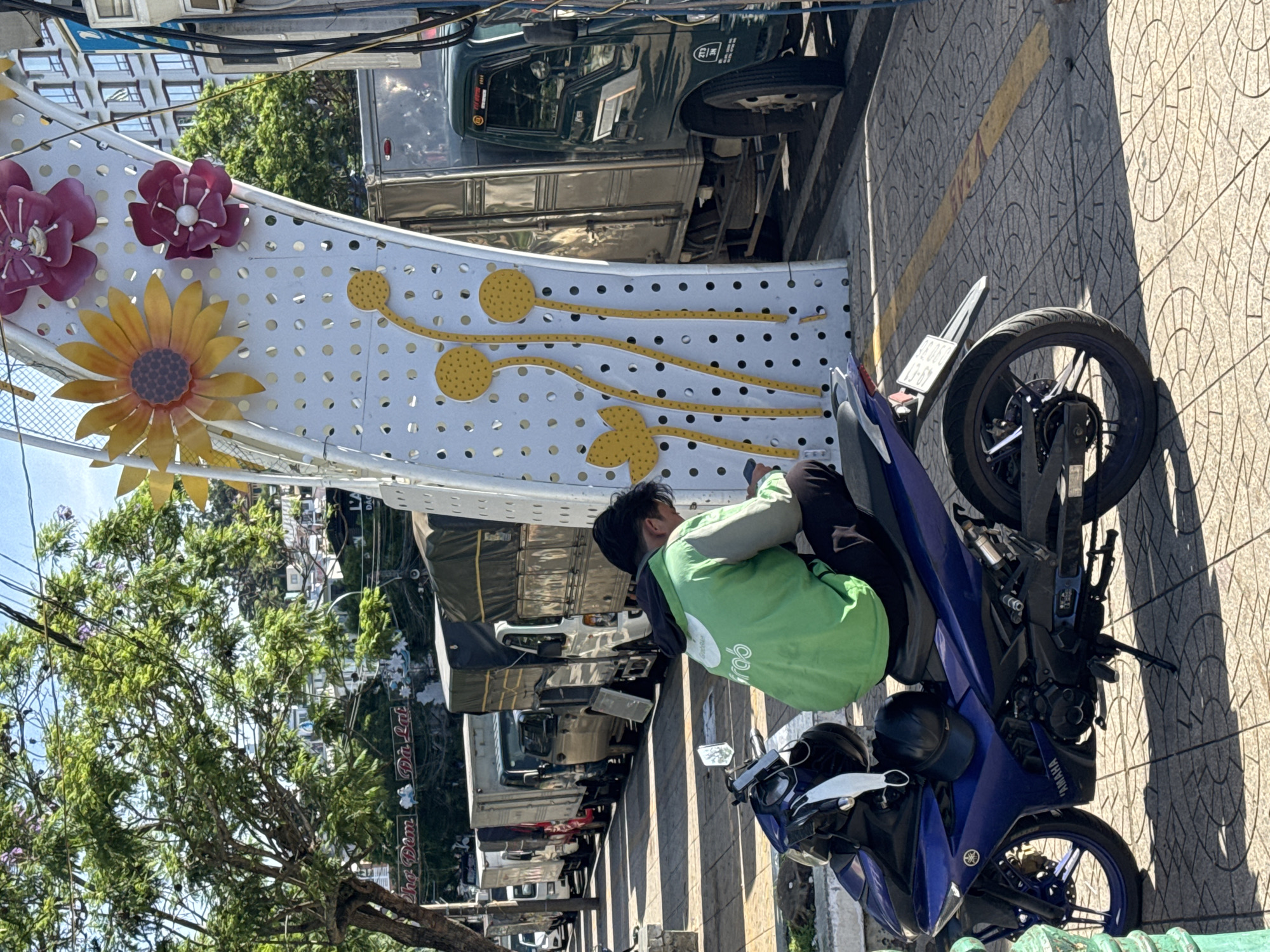 A young man wearing a bright green hooded jacket with a "Grab" logo is crouched on a blue motorcycle, looking at his phone. He is parked on a tiled sidewalk in Da Lat, Vietnam, next to a large, white arched structure decorated with large yellow and pink artificial flowers and yellow lotus-like motifs. Behind the man and the structure is a line of parked trucks. The environment is sunny with shadows cast on the ground. The background shows trees and buildings.