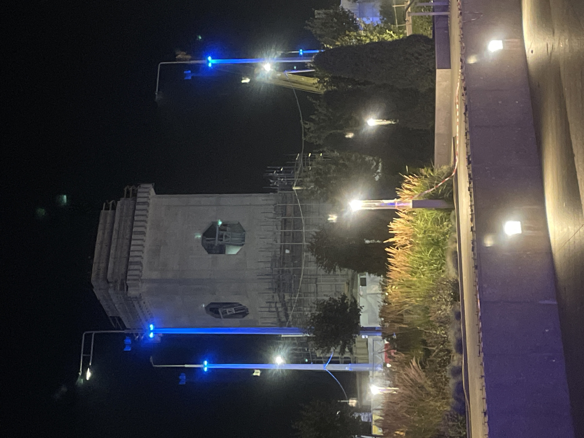 The image shows a nighttime scene in Saint Petersburg, Russia, likely near the Zaryadye Park, featuring a partially constructed tower illuminated by blue lights. The tower, made of concrete and surrounded by scaffolding, has octagonal openings. Several streetlights with bright white lights are visible, along with decorative lighting at the base of the tower and along a walkway. In the foreground, there is a landscaped area with tall, dry grasses and decorative shrubs. The ground in the foreground is paved, and a warning tape is visible across the path. The overall impression is of an urban construction site at night, with lighting creating a dramatic effect. There are no people visible in the image.