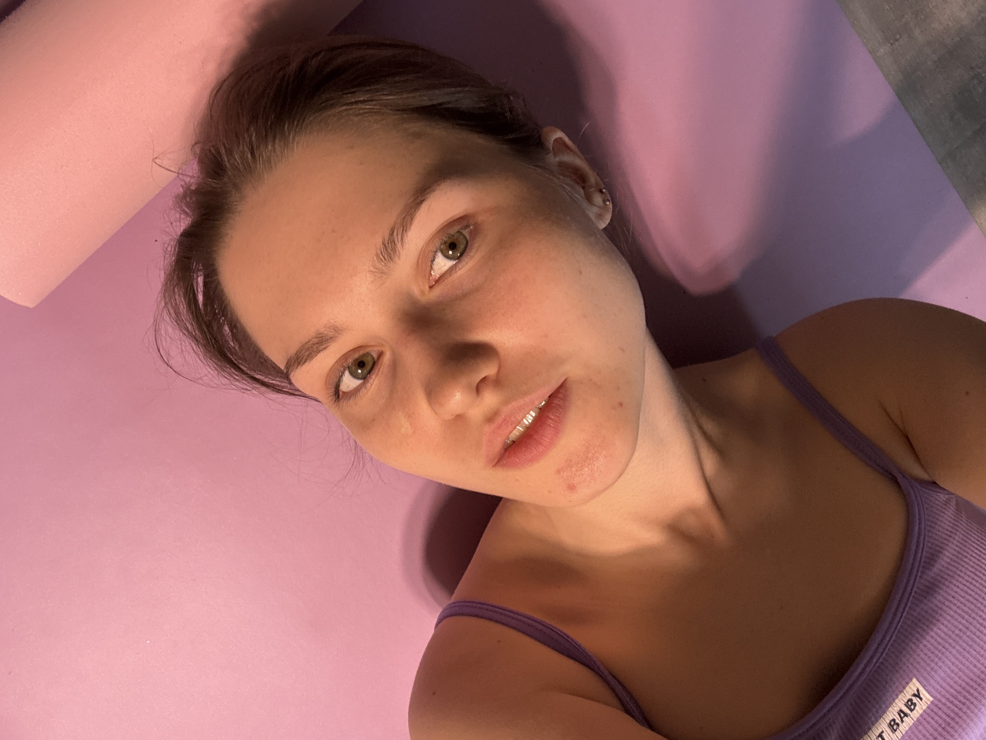 A woman lies on a purple yoga mat, wearing a lavender tank top with "SWEET BABY" printed on it. The lighting is diffused and slightly pink-toned. The background shows a pink foam roller and a section of gray flooring. The woman is fair-skinned with light brown hair pulled back from her face, and she has green eyes. She appears to be looking at the camera. The location is not discernible from the image.