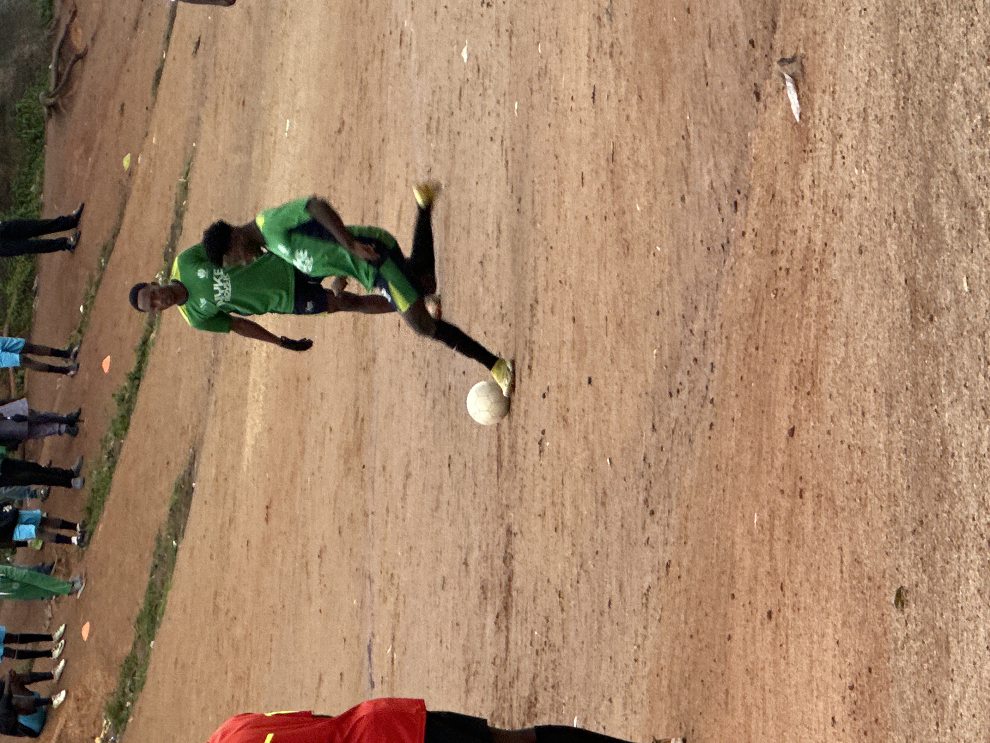 The image depicts a dirt soccer field in Jos, Nigeria, with several individuals in athletic wear. In the foreground, a person wearing a red shirt and black shorts is partially visible on the left. The central focus is a soccer player in a green jersey and black shorts, with yellow cleats, kicking a white soccer ball. Another player in a similar green jersey stands behind him, observing. Other individuals in athletic attire are scattered in the background, some seated and others standing, on a slightly elevated section of the field. The environment is characterized by a dusty, reddish-brown dirt surface with sparse patches of green vegetation along the edges. The overall scene suggests an informal soccer training or practice session.