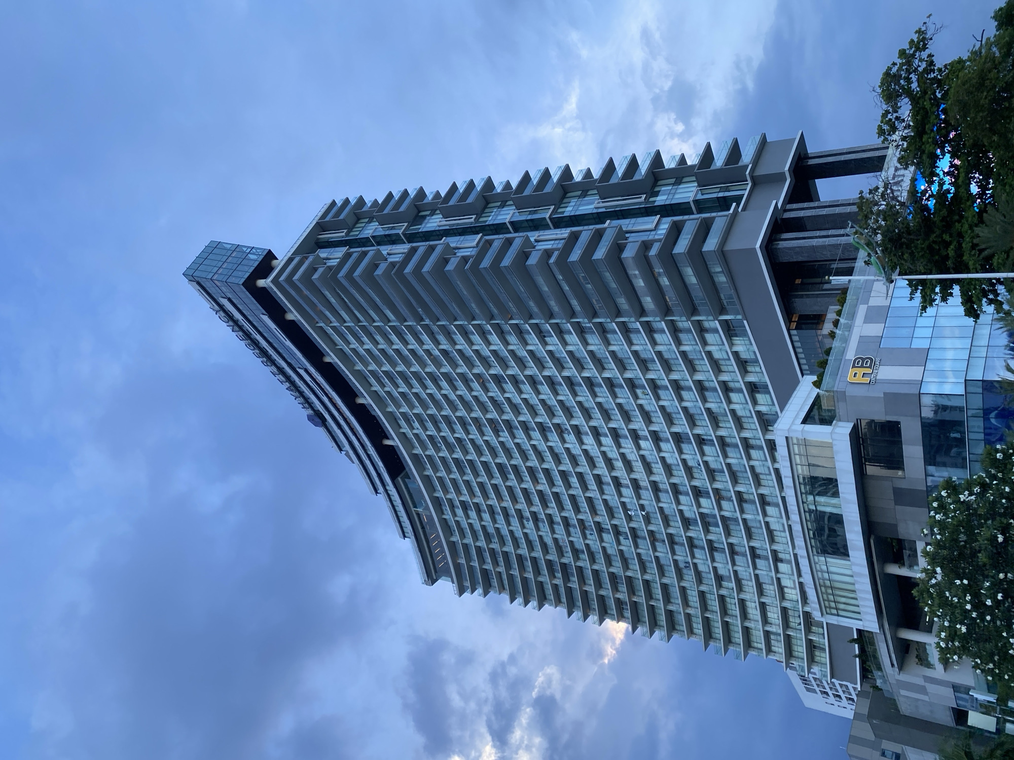 This media file shows a modern, tall building with a curved facade, likely a hotel or residential complex, viewed from a low angle against a cloudy sky. The building has numerous balconies or recessed windows that create a layered, stepped effect. The structure is predominantly gray and glass, with some darker sections. On the lower part of the building, a glass facade reflects the sky and surrounding trees. A logo with the letters "BP" inside a yellow square and the text "CENTRAL SQUARE" is visible on the lower section of the building. To the right, lush green trees with white flowers partially obscure the building. The lighting suggests it is late afternoon or early evening, with overcast weather. The scene is static, with no people or activities depicted, focusing solely on the architecture and its surroundings. The location context provided is Nha Trang Ward, Vietnam.
