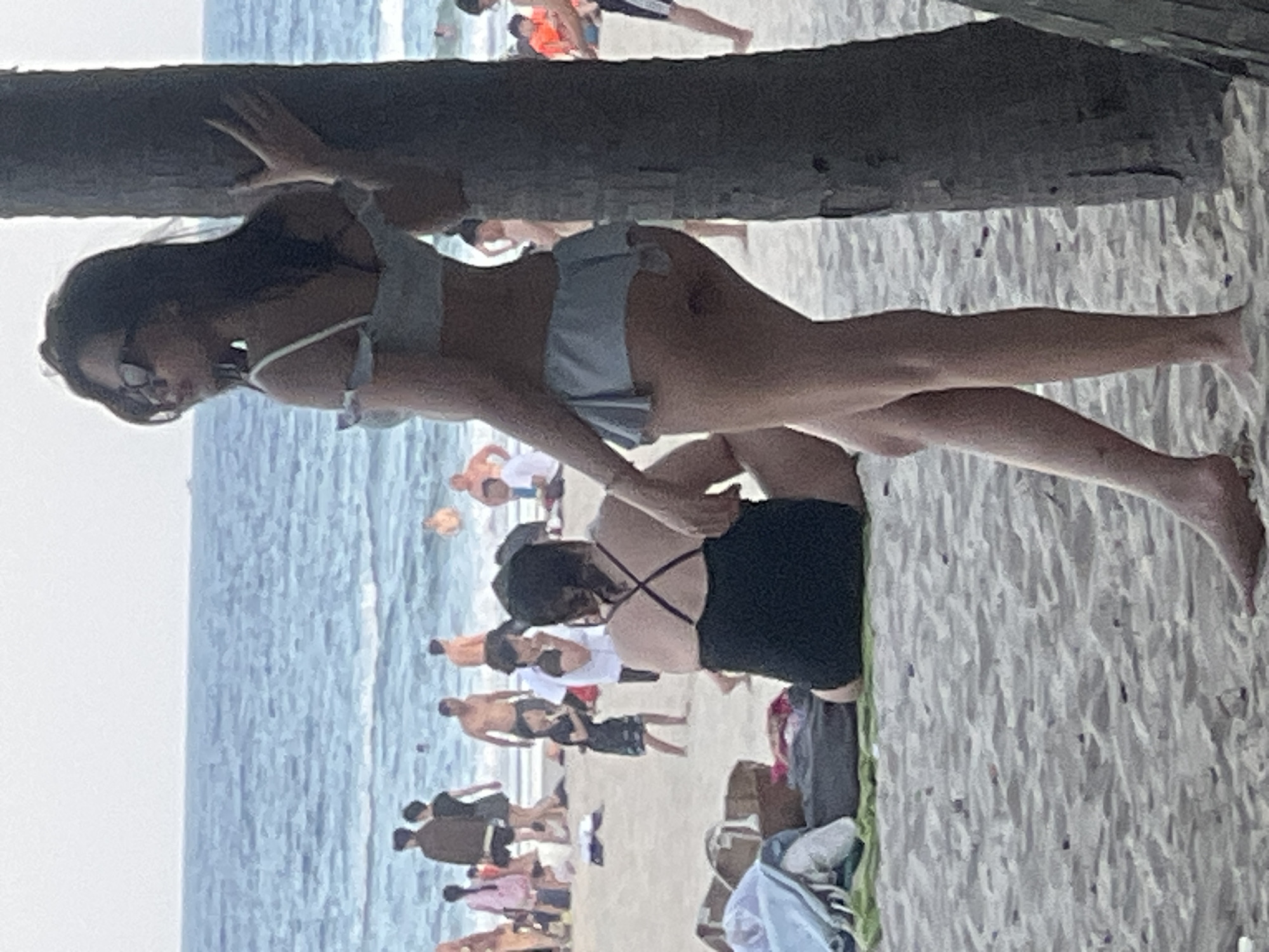 In this image, two women are on a beach in Đà Nẵng, Vietnam, near the ocean. The woman in the foreground is leaning against a palm tree, wearing sunglasses and a light-colored bikini. Her body is positioned with her legs extended. Behind her, another woman is sitting down, wearing a black swimsuit. The beach is populated with other people, some walking, others relaxing, and children are visible in the background. The water appears calm, and the sky is bright, suggesting it is daytime with fair weather. The presence of palm trees and the general beach atmosphere are strong indicators of the tropical coastal setting of Đà Nẵng. There is no visible text in the image.