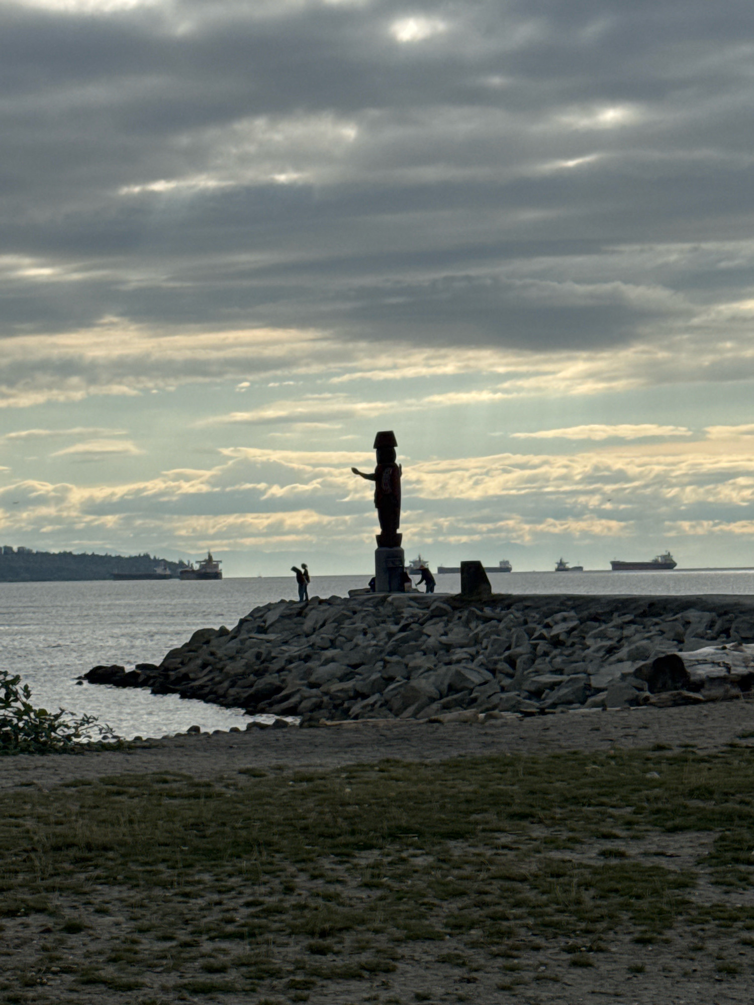The image captures a view of the ocean with a rocky shoreline in the foreground. A totem pole stands on the rocks, silhouetted against a cloudy sky. Several cargo ships are visible in the distance, moving across the water. A few people are standing on the rocks near the totem pole. The shoreline transitions to a grassy area in the lower portion of the image. The scene appears to be located in West Vancouver, Canada, possibly near Brockton Point, a known location for totem poles and views of the Vancouver harbour. The overall atmosphere is serene and somewhat dramatic due to the overcast sky and the silhouettes.