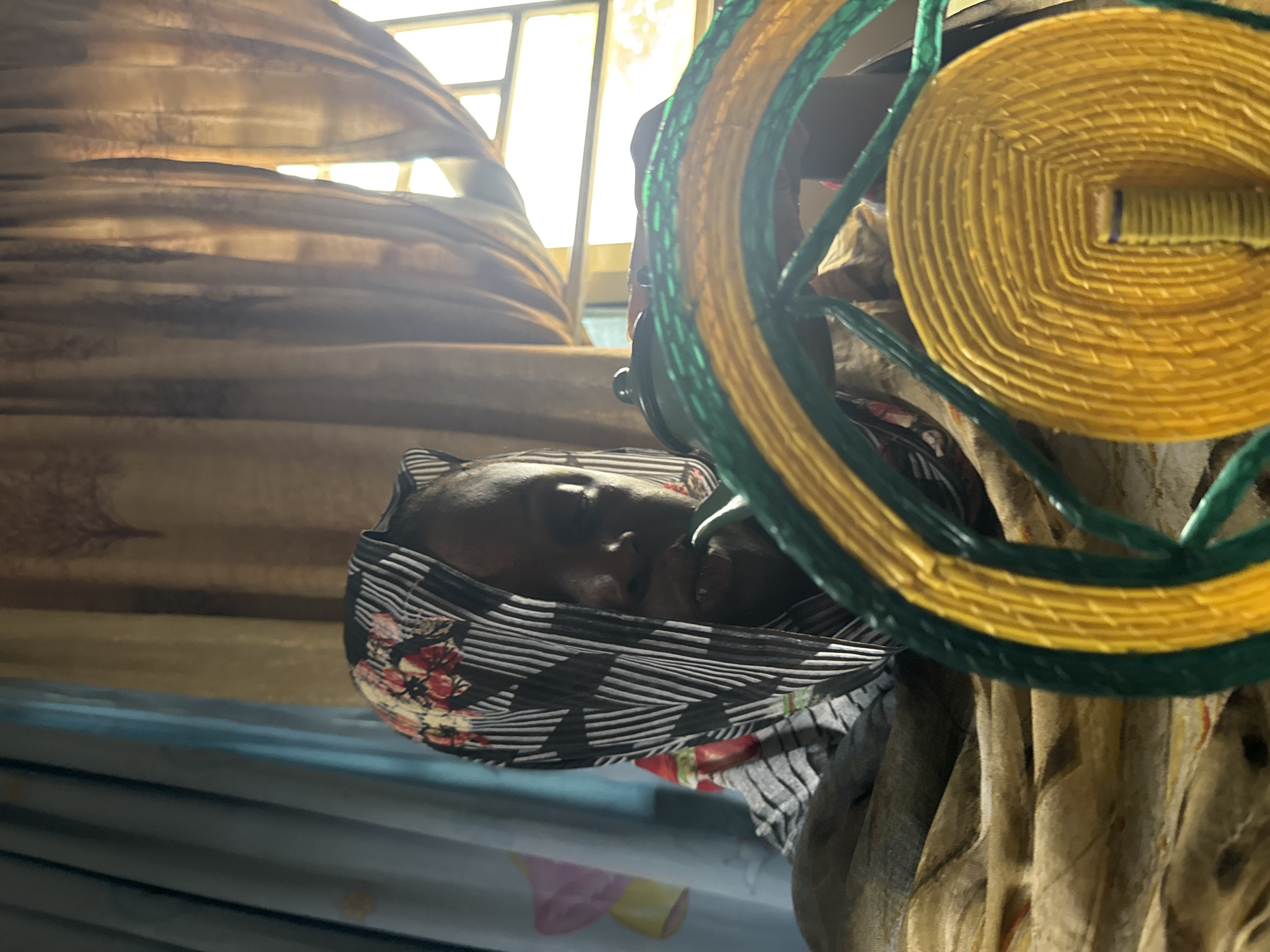 A person with a headscarf, lying down on a blue surface with a yellow and green woven circular object. The person is likely indoors in a dwelling with window curtains. The location is Madangala, Nigeria.