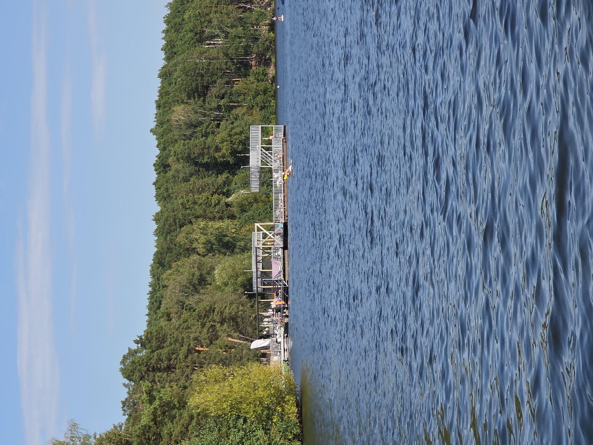 A sunny day at a lakeside recreational area is depicted. The foreground is dominated by rippling blue water, likely reflecting the clear sky. In the middle ground, a long, white, multi-level wooden pier extends into the water. The pier has railings, stairs, and several people on it, some appearing to be sunbathing or socializing. A large white umbrella-like structure is visible on the pier. To the left of the pier, a small white boat is docked. Further out on the water, two individuals are on paddleboards, one of whom is actively paddling. In the background, a dense forest of green pine trees lines the shore, with a few lighter-colored deciduous trees interspersed. The sky above is a bright, clear blue with a few wispy white clouds. This scene is consistent with a view of the embankment or a recreational zone along Lake Chebarkul in Chebarkul, Russia, a known resort area.