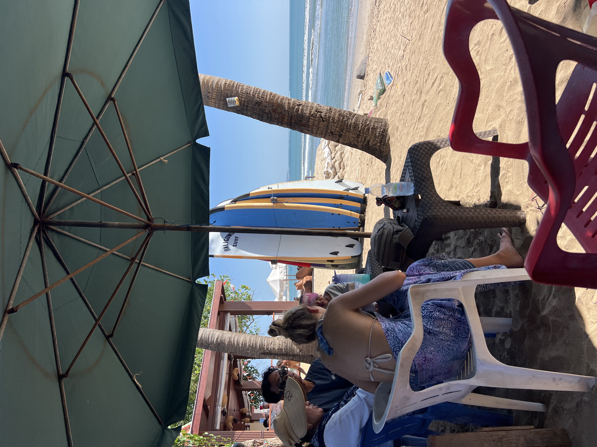 An eye-level, medium shot captures a beachside dining scene in Kuta, Indonesia, under the shade of a large green umbrella. Four individuals are seated around a woven plastic table on a sandy beach. The person closest to the viewer, wearing a bikini top and a purple sarong, is seen from behind. To their left, a person in a white shirt and a straw hat faces them. Another individual in a dark shirt and sunglasses is seated to the left, and a fourth person, partially obscured, is seated across from the person in the sarong.
On the table, a bottle of "Le Minerale" water and a dark bag are visible. Behind the table, two surfboards, one white with blue and yellow accents and the other predominantly blue, lean against a palm tree. In the background, the turquoise ocean stretches to the horizon, with gentle waves rolling onto the shore. The beach is dotted with sandbags and a few stray items, suggesting a relaxed, tropical atmosphere. The lighting indicates it is daytime.