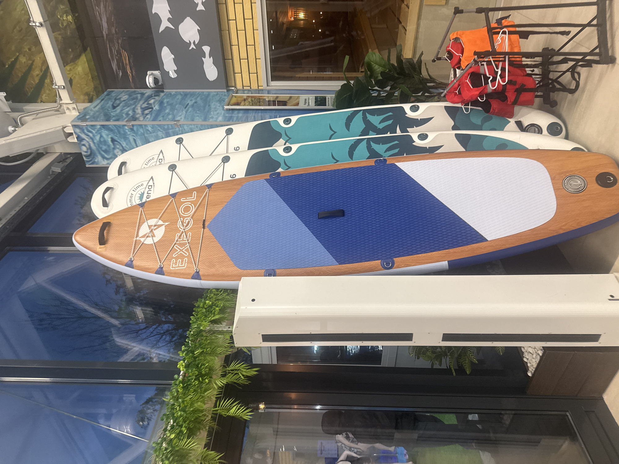 The image shows a display of paddleboards outside a building. Three paddleboards are stacked horizontally. The top board is mostly blue and white with a wood-grain colored trim. The word "EXEGOL" is printed on it. The other two boards have a white base with teal and green accents. Life vests hang on a metal rack behind the boards. The location cannot be verified as Ulyanovsk, Russia.