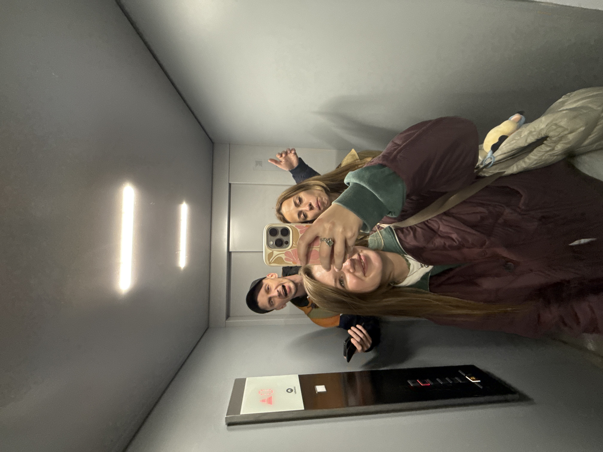 The image shows a mirror selfie taken by three individuals inside a modern elevator. The main subject, a woman with long, light brown hair, is in the foreground on the right, holding a smartphone with a floral-patterned case. She is smiling broadly at the camera. Behind her, slightly to the left, is a man wearing a dark beanie and a striped top, also smiling with his mouth open. Further back and to the left, another woman is visible, smiling and making a gesture with her hand, possibly a peace sign.
The elevator interior features light grey walls and a white ceiling. Bright, vertical strip lights illuminate the left wall. At the bottom of the image, the elevator control panel is visible, made of metallic and black sections. A red illuminated "10" is clearly displayed, likely indicating the current floor, along with an illuminated upward arrow. Other buttons and symbols are present but less distinct. A small, light blue and yellow plush toy is clipped to a bag or jacket in the upper right quadrant of the image. The subjects appear joyful and are actively posing for the photograph.