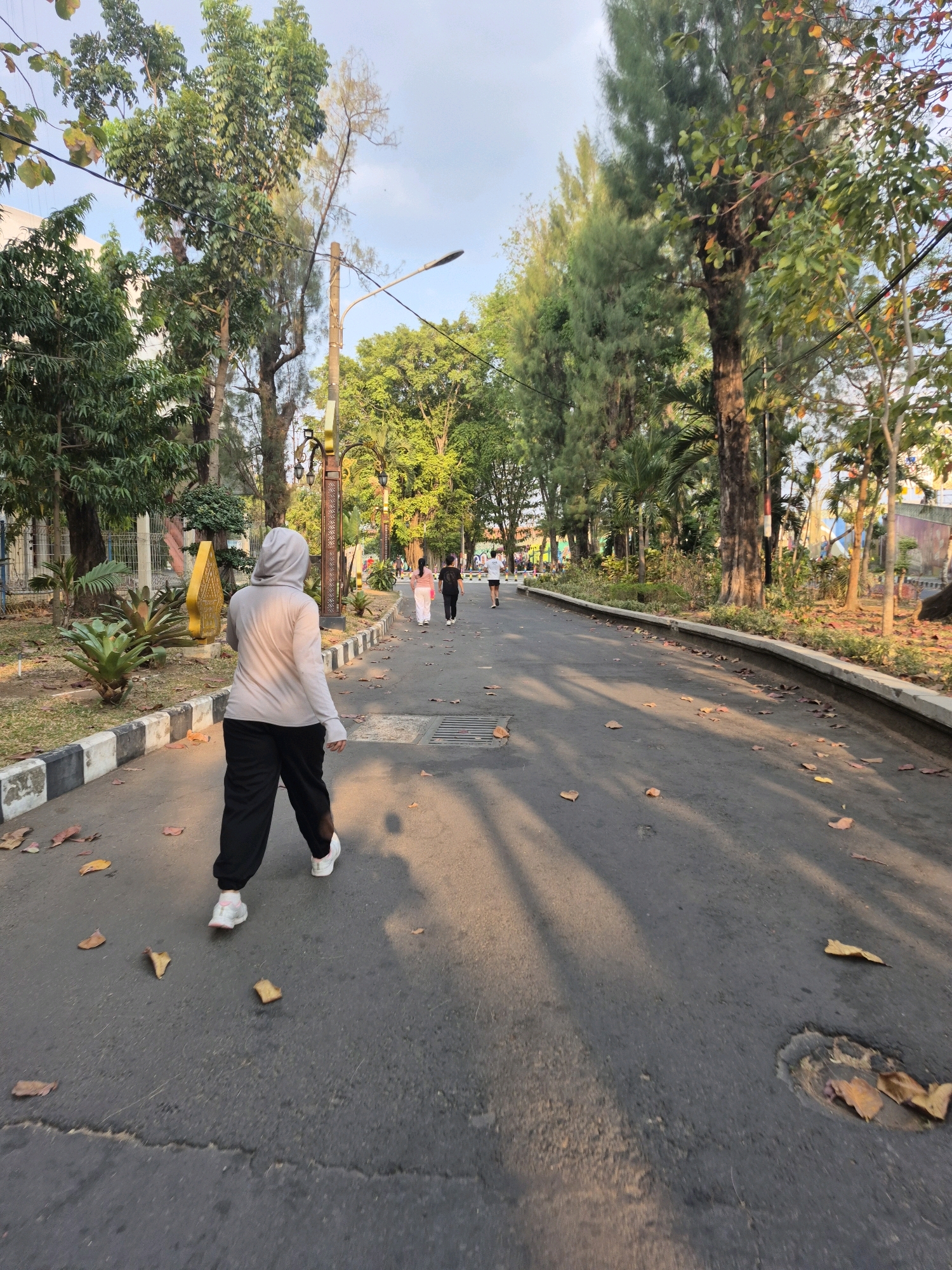 An outdoor, medium-shot, eye-level, full-body perspective of a young woman wearing a light gray hijab, a light beige long-sleeved t-shirt, and black pants walks away from the camera on a paved path in a park in Surakarta, Indonesia. Her arms are at her sides, and her head is turned slightly to her right. She wears white sneakers with pink accents. In the middle ground, three people walk in the same direction: a woman in a pink shirt and white pants, a woman in a white shirt and dark pants, and another person in dark clothing. The path is lined with short, black-and-white striped borders, followed by grass and trees. Large, leafy trees with green and brown foliage line both sides of the path, with some fallen leaves scattered on the ground. A streetlamp is visible in the distance to the left, and a decorative golden structure stands near the path on the left side. The sky is a light blue with some clouds. The sun casts long shadows from the trees across the path, indicating late afternoon or early morning.