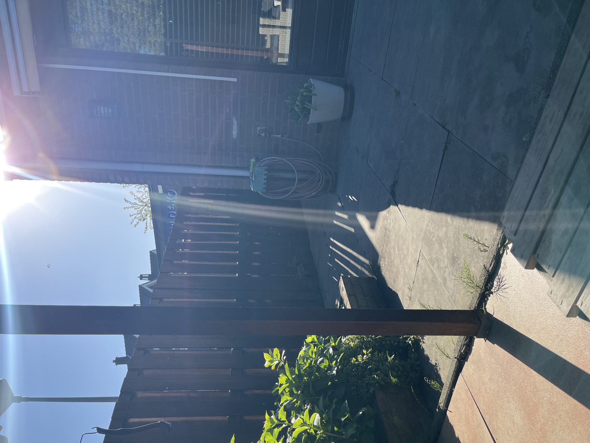 The image displays a sun-drenched residential backyard or patio, captured from a slightly elevated vantage point. Intense sunlight pours in from the left, creating a prominent lens flare and stark contrasts between brightly illuminated areas and deep shadows cast across the scene.
On the left side, the exterior of a brick house is visible, featuring a large window with horizontal blinds and a small rectangular vent. Below the window, a garden hose is coiled on a wall-mounted reel, positioned next to a water spigot. A large white pot, containing vibrant green foliage, rests on the paved ground. The ground is covered with grey paving slabs, where some weeds have grown through the cracks.
A wooden fence or structure, made of vertical posts and horizontal slats, stretches across the middle ground, casting long, distinct shadows on the patio. Beyond the fence, leafy green plants flourish in garden beds, and a small, deciduous tree is silhouetted against a clear, bright blue sky. Two small birds are visible, flying high in the sky. Reflections of interior elements are discernible in the house window. Behind the fence, a blue object with white capital letters, possibly spelling "OTEK" or "OTECO," is partially visible. The scene suggests a clear, sunny day, with the low-angle sun indicating either early morning or late afternoon. The overall atmosphere is serene and domestic.