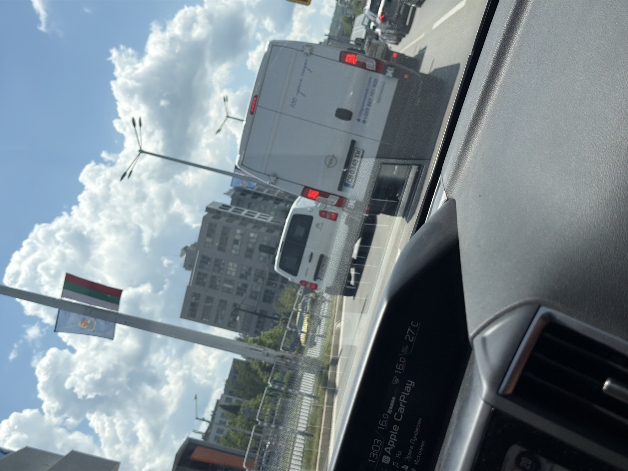 The image shows a view from inside a vehicle, looking out at traffic on a sunny day in Sofia, Bulgaria. The dashboard of the vehicle displays "Apple CarPlay," the time "13:03," a temperature of "16.0" degrees Celsius, and an outside temperature of "27°C." Several vehicles are visible on the road, including a white van with the license plate "CB 0348 XM" and text on its rear that includes a phone number. In the background, multi-story buildings and streetlights can be seen. A Bulgarian flag and another flag are visible on a flagpole. The sky is blue with scattered white clouds.