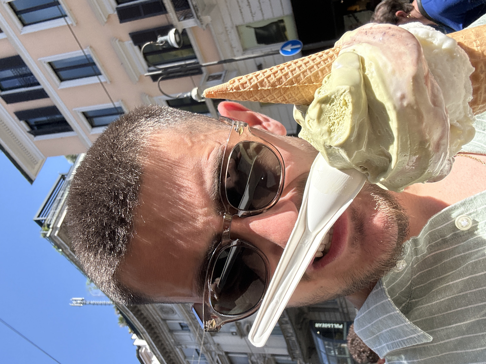 This vibrant, rotated close-up captures a male subject enjoying a large ice cream cone outdoors on a sunny day in Milan, Italy. The man, with short dark hair and stubble, is depicted from the chest up, wearing sunglasses with dark lenses and a light green and white striped collared shirt. He is holding a generously scooped ice cream cone, which appears to feature at least three flavors: a prominent green (likely pistachio), white (possibly vanilla or cream), and a reddish-pink swirl. A white plastic spoon, subtly marked with "RST", is positioned in his open mouth, conveying an expression of enjoyment, possibly a smile or a laugh, as he consumes the ice cream.
The background shows an urban environment under a clear blue sky. Behind the man, a multi-story building with a light pink/beige facade, distinct rectangular windows with dark frames, and white architectural details is visible. Further into the background, other urban structures and a blue directional arrow sign on a pole can be discerned. A reverse reflection of the "PULL&BEAR" store name is also visible on a window. The strong sunlight casts shadows, indicating a bright daytime setting. The photograph is taken from a low-angle perspective, looking upwards, and is presented rotated 90 degrees counter-clockwise.