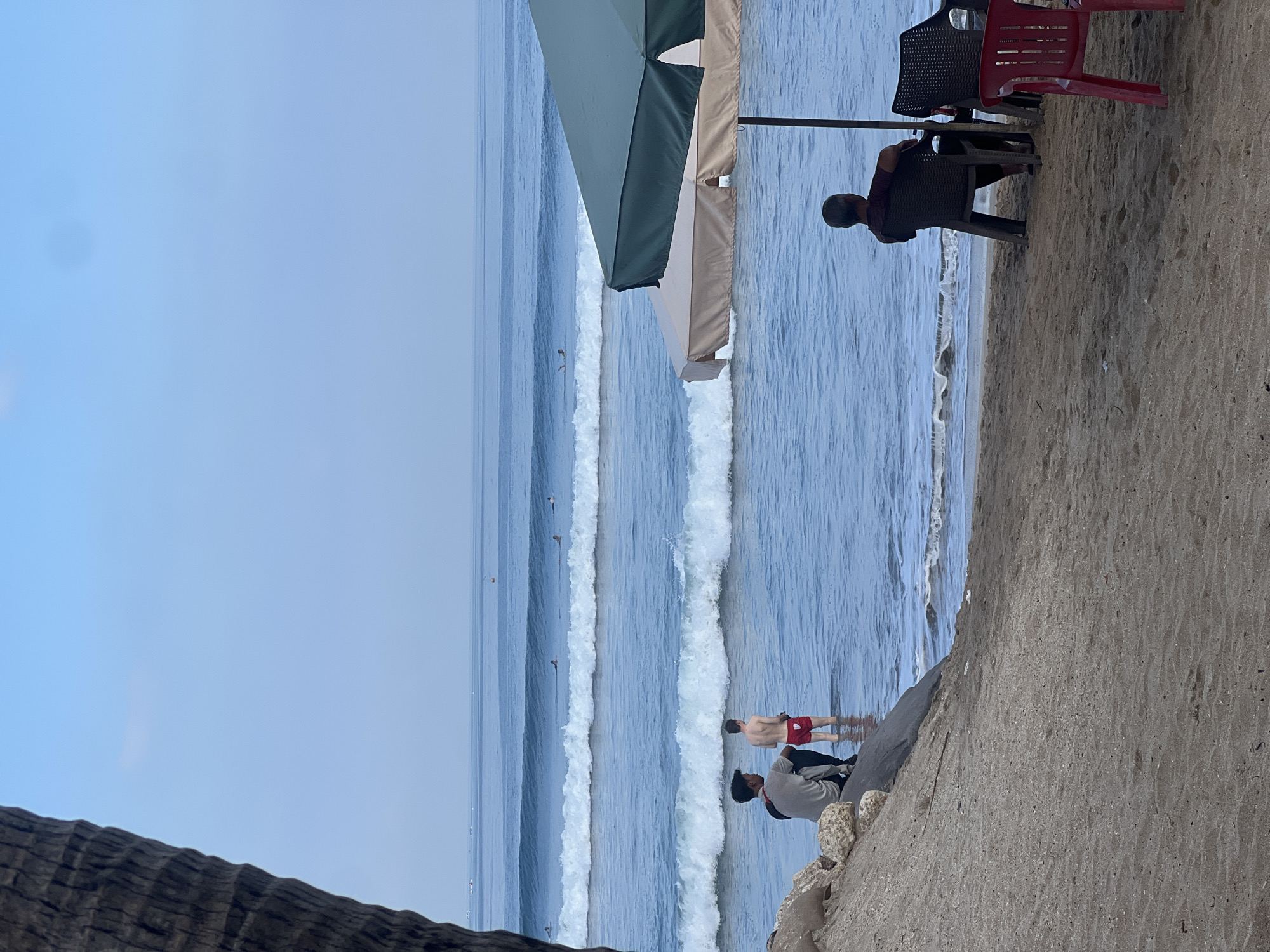 The image captures a serene beach scene in Kuta, Indonesia. In the foreground, a person sits on a sandy beach under a large umbrella, facing the ocean. Two other individuals are partially visible near the water's edge, one standing and the other sitting. Several surfers are dotted across the calm, blue ocean, with multiple waves rolling towards the shore. The sky is clear and a pale blue, suggesting it might be early morning or late afternoon. A large palm tree trunk is visible on the left side of the frame, adding a tropical element to the setting. The beach is composed of fine, light-colored sand, with some rocks and debris scattered near the waterline. The overall atmosphere is peaceful and relaxed, typical of a popular beach destination like Kuta.