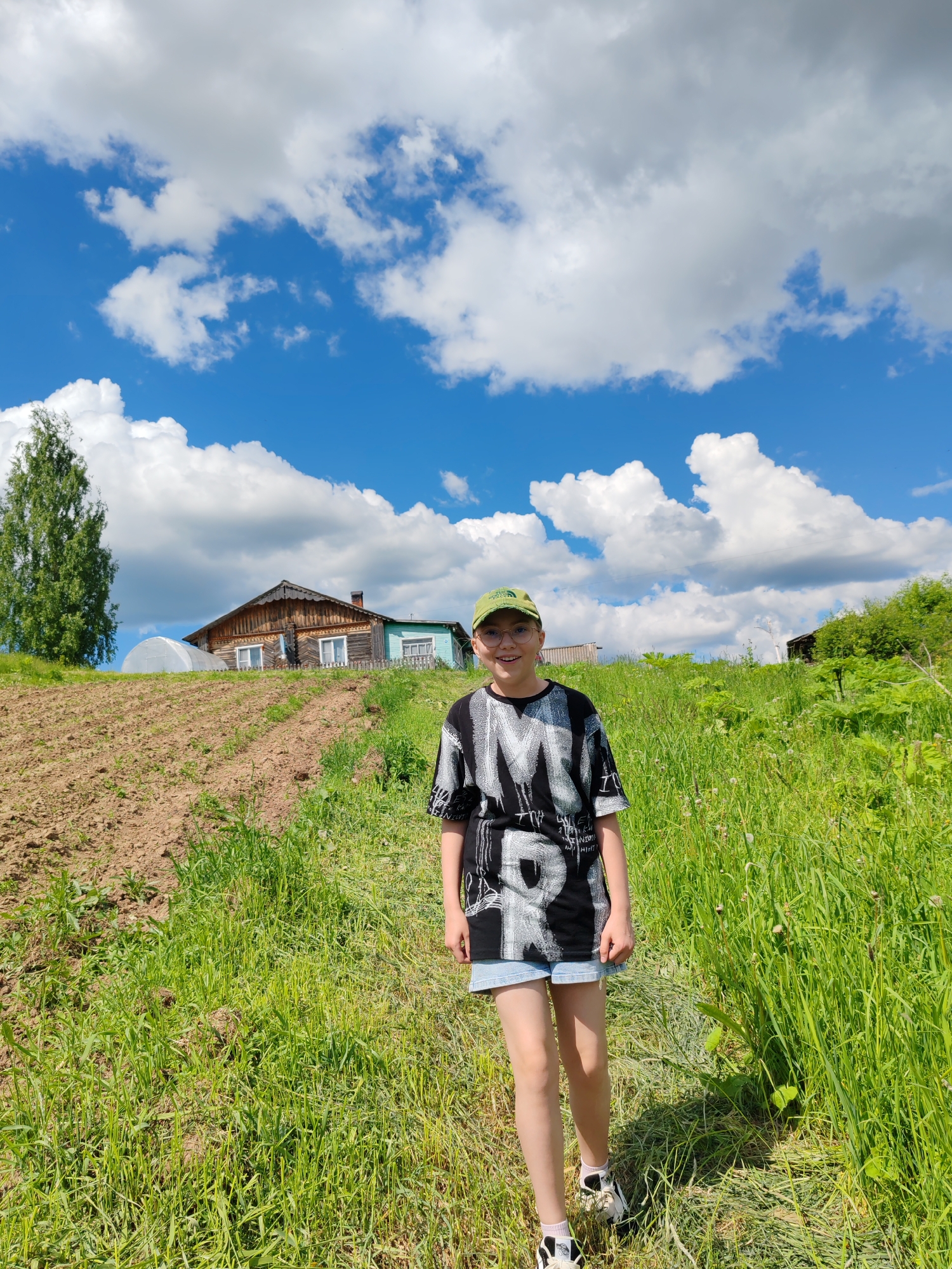 The image depicts a rural outdoor scene in Чухлэм, Russia, under a clear blue sky with substantial white cumulus clouds. A young individual stands centrally in the foreground, facing slightly right of the camera with a subtle smile. The person wears a green baseball cap, round glasses, a black short-sleeved t-shirt with prominent white graphic lettering, light blue denim shorts, white socks, and black-and-white athletic shoes.
The immediate foreground consists of dense, bright green grass mixed with some small plant growth, forming a narrow, cleared path where the person stands. To the left of the person is a section of recently tilled, dark brown earth, sloping upwards. Further left, a tall, slender deciduous tree with vibrant green foliage is visible.
In the midground, situated on a gentle incline, is a traditional wooden house. The house features a weathered brown wooden facade, white-framed windows, intricate detailing along the roofline, and a visible chimney. An extension or adjacent structure, painted a lighter blue, is attached to the right side of the main house. To the left of the house, a white, semi-circular greenhouse structure is present. The background to the right shows additional green foliage and subtle hints of other distant structures. The scene is brightly illuminated by natural sunlight, indicating a clear day.