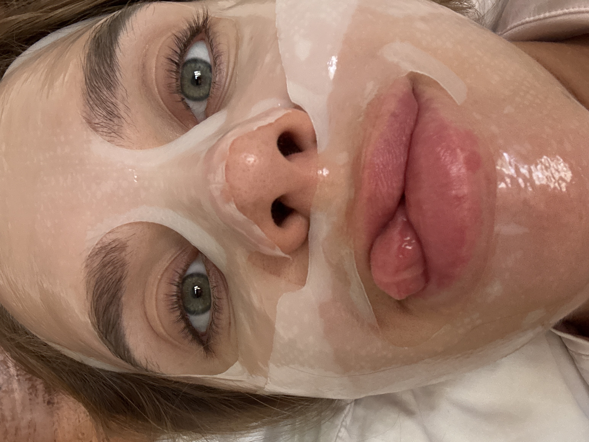 A close-up, high-angle shot depicts a person wearing a transparent facial sheet mask. The mask covers most of the face, conforming to the contours of the forehead, cheeks, nose, and chin. The lips are uncovered and appear slightly puckered. The person's eyes, framed by eyelashes and eyebrows, are visible through the mask's openings. A portion of the person's hair is visible on the side of the face. The subject is situated in North Kuta, Indonesia.