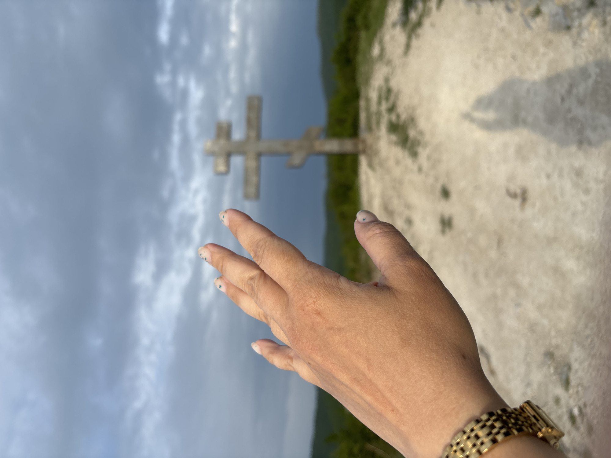 A low-angle shot features a person's extended hand with painted fingernails and a gold watch on their wrist. In the background, a stone Christian cross stands on a hill against a cloudy sky in Semogorskiy, Russia. The ground is light-colored, possibly gravel. Some green vegetation is visible in the background.