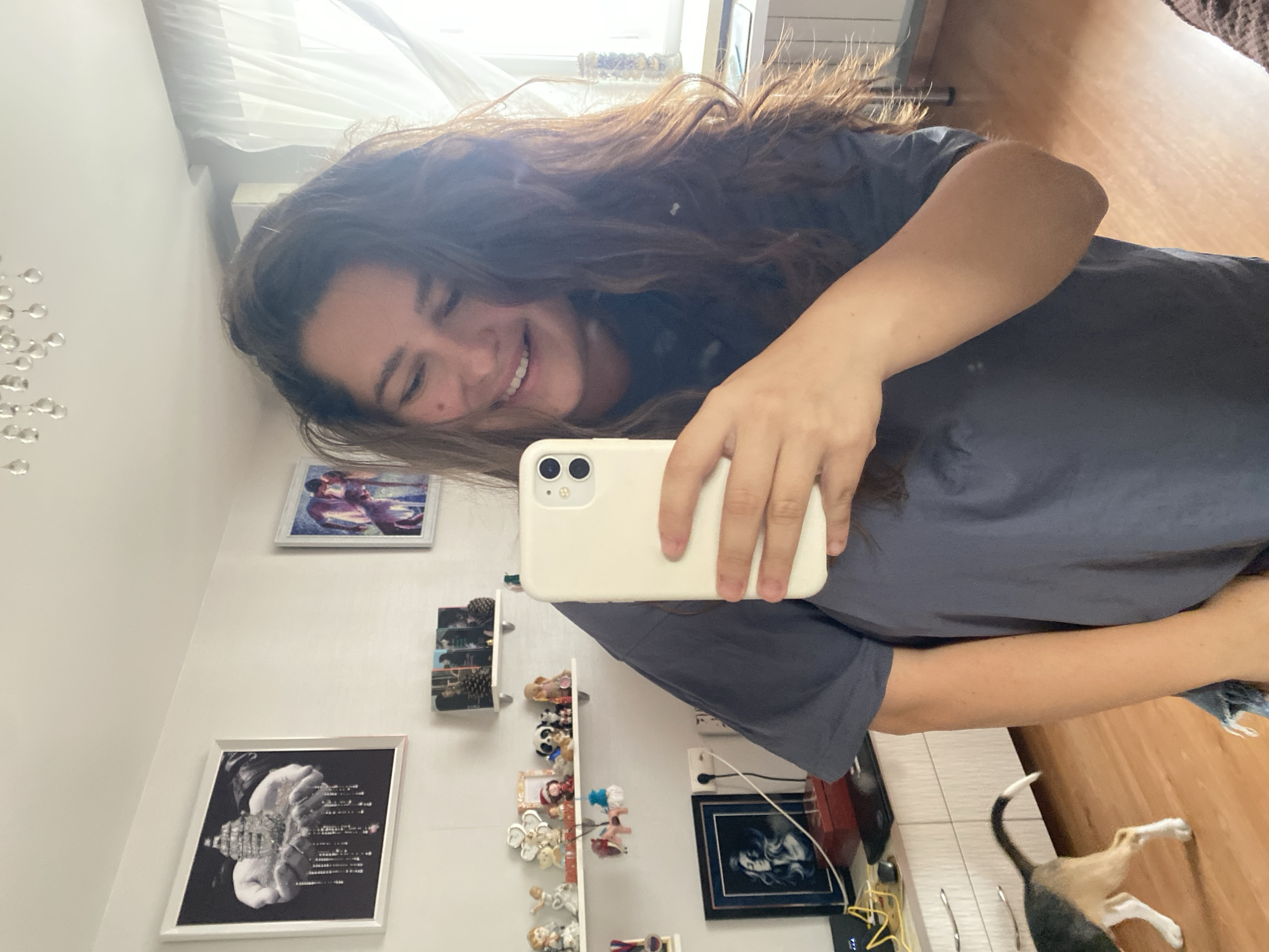 The image shows a young woman taking a selfie in a room. She has long, dark, wavy hair and is smiling. She is wearing a dark t-shirt. The room appears to be a living space, with several framed pictures on the wall, including a black and white one and a colorful one. There are also shelves filled with small decorative items and figurines. A white iPhone is held in the woman's hand, and a cat can be seen in the lower right corner of the image. The lighting suggests it could be daytime. The image is oriented vertically, and the overall mood is casual and happy.
