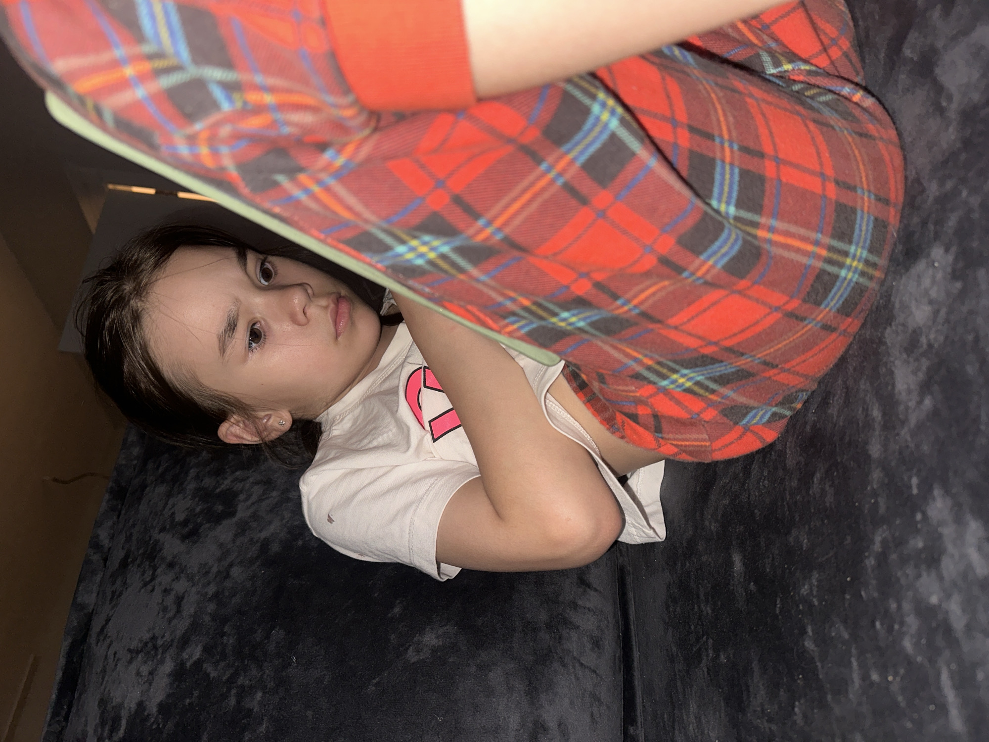 A girl is lying on a dark gray couch. She is wearing a white t-shirt and red plaid pajama pants with a blue and yellow grid pattern. The location is unknown; the claim about Kharnavy, Russia is not verifiable from the image.
