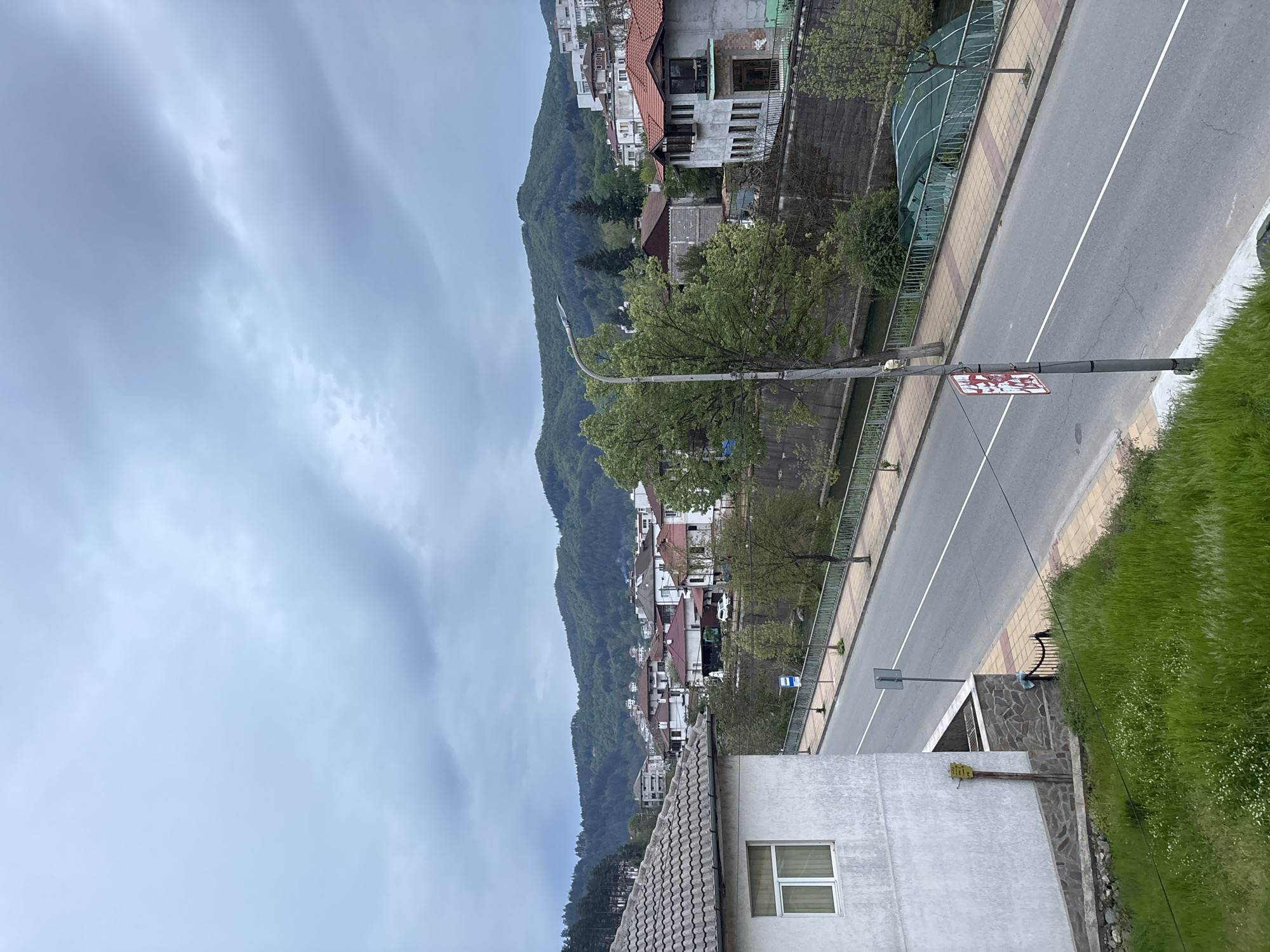 The image captures an elevated, slightly rotated view of Zlatograd, Bulgaria. A gray, overcast sky dominates the top left. In the middle ground, a mountain is covered with a dense forest, with a line of houses visible along its base. A road runs parallel to the mountain, separated by a fence. A white line marks the center of the road. A light post stands on the side of the road, with a sign affixed to it. A white building with a gray tiled roof and a single visible window is in the foreground. Green vegetation is on the right side of the road.