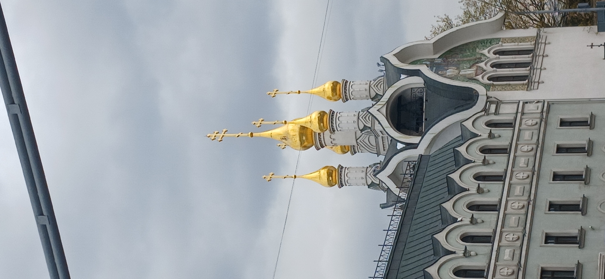 An architectural photograph depicts a grand building, likely a Russian Orthodox church, characterized by its distinctive golden onion domes, set against an overcast sky. The image is oriented sideways, but if viewed upright, the building stands prominently on the right.
The central feature is a section of the building topped with three prominent golden onion domes, each crowned with an Orthodox cross. These domes sit atop white, multi-tiered drum structures with intricate grey and white decorative patterns. Beneath the domes, the roofline is dark grey with a scalloped, ornate edge.
To the right of the domed section, an adjacent building or wing features a lighter facade with multiple rows of rectangular windows. This section also incorporates decorative elements, including a continuous band of arched windows or alcoves along its upper part and what appears to be a mosaic or fresco depicting figures on its uppermost level, partially obscured by tree branches. The lower part of this section has a series of small, dark rectangular windows.
The sky is a uniform, light grey, indicating an overcast day. On the far left of the frame, a dark grey, vertical pole or beam is partially visible, extending from bottom to top. The overall impression is one of traditional Russian ecclesiastical architecture, consistent with the provided location context of Moscow, Russia. There is no visible text in the image.