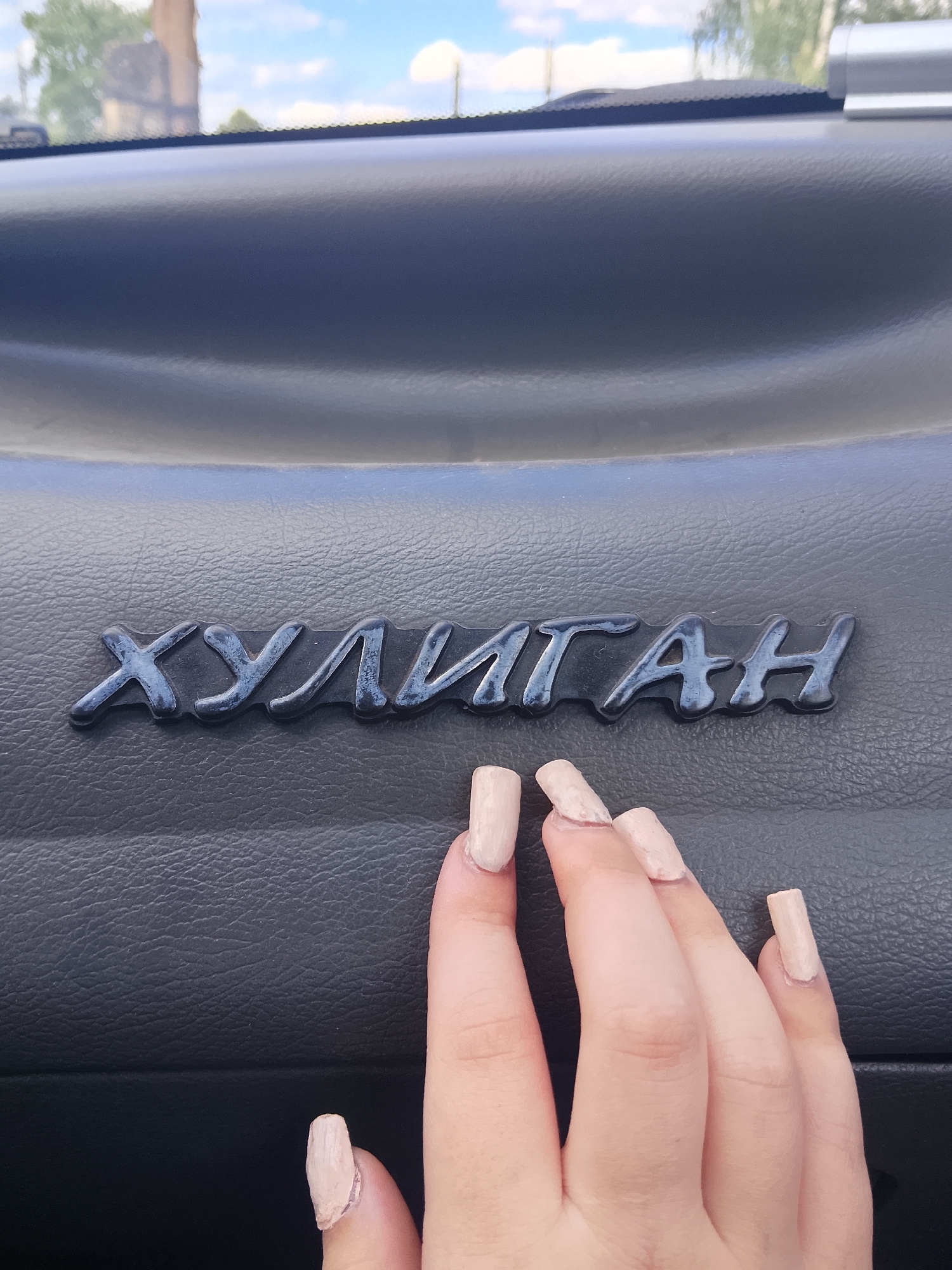 A close-up, eye-level shot shows a black emblem on a textured black surface, likely a car dashboard, in Verkhopol'e, Russia. The emblem consists of cursive Cyrillic letters that spell out "ХУЛИГАН" (KHULIGAN). The letters are glossy black with some blue and white highlights, suggesting they are metallic or painted. The textured surface beneath the emblem has a matte finish. In the foreground, a person's hand with light-colored nail polish and long fingernails is positioned partially over the emblem and the lower portion of the dashboard. The background, visible through a windshield or window, shows an outdoor scene with a cloudy blue sky and hints of greenery and a building, indicating a daytime setting. The lighting suggests natural daylight.