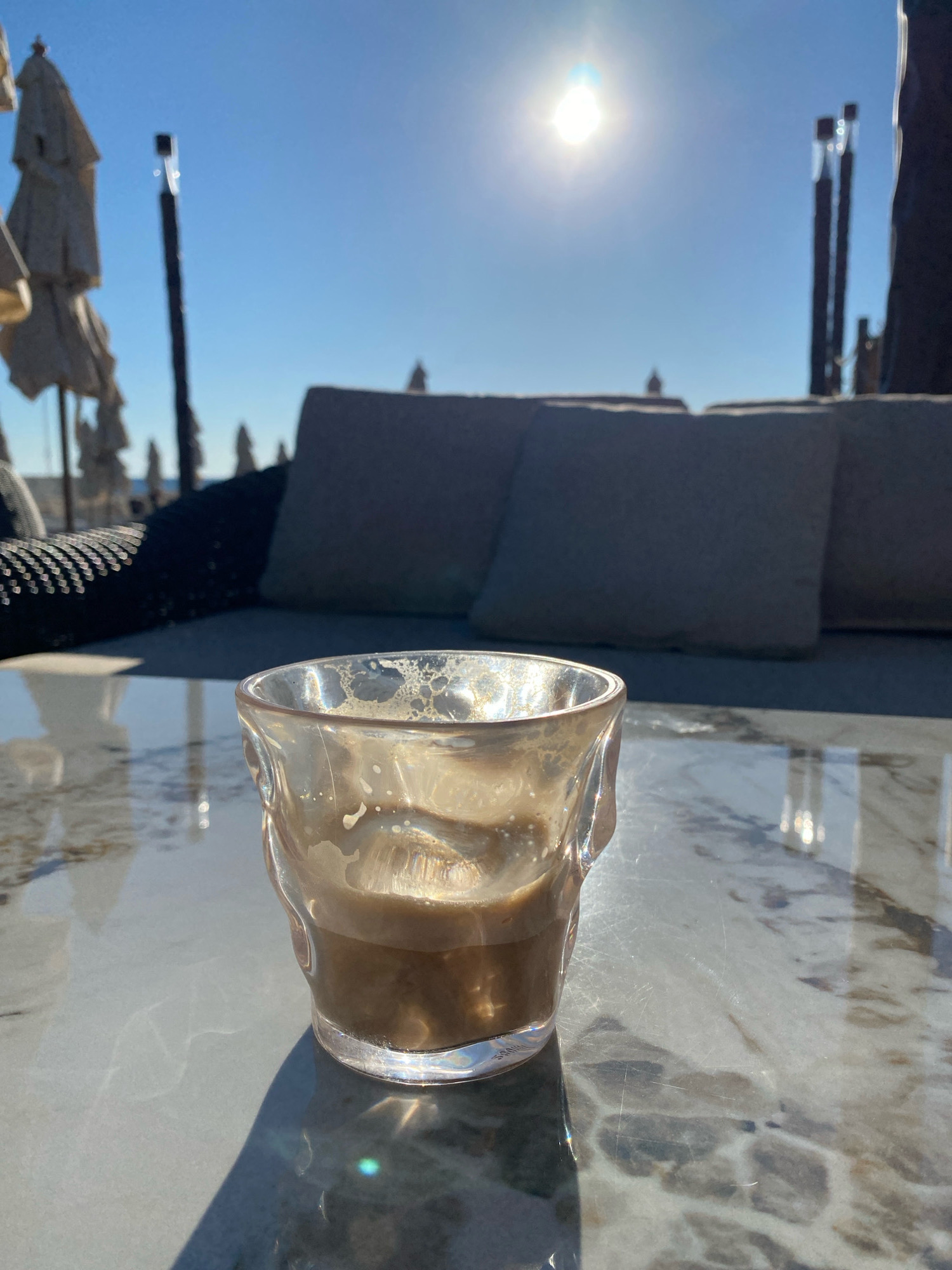 A glass with a small amount of coffee and foam is positioned on a marble table. The table reflects the bright sunlight and the surrounding environment. In the background, several folded beach umbrellas are visible, along with cushioned seating and what appear to be thin poles. The sky is clear and blue with a bright sun at its zenith. This scene suggests an outdoor seating area, possibly at a cafe or resort, in Odesa, Ukraine, on a sunny day.