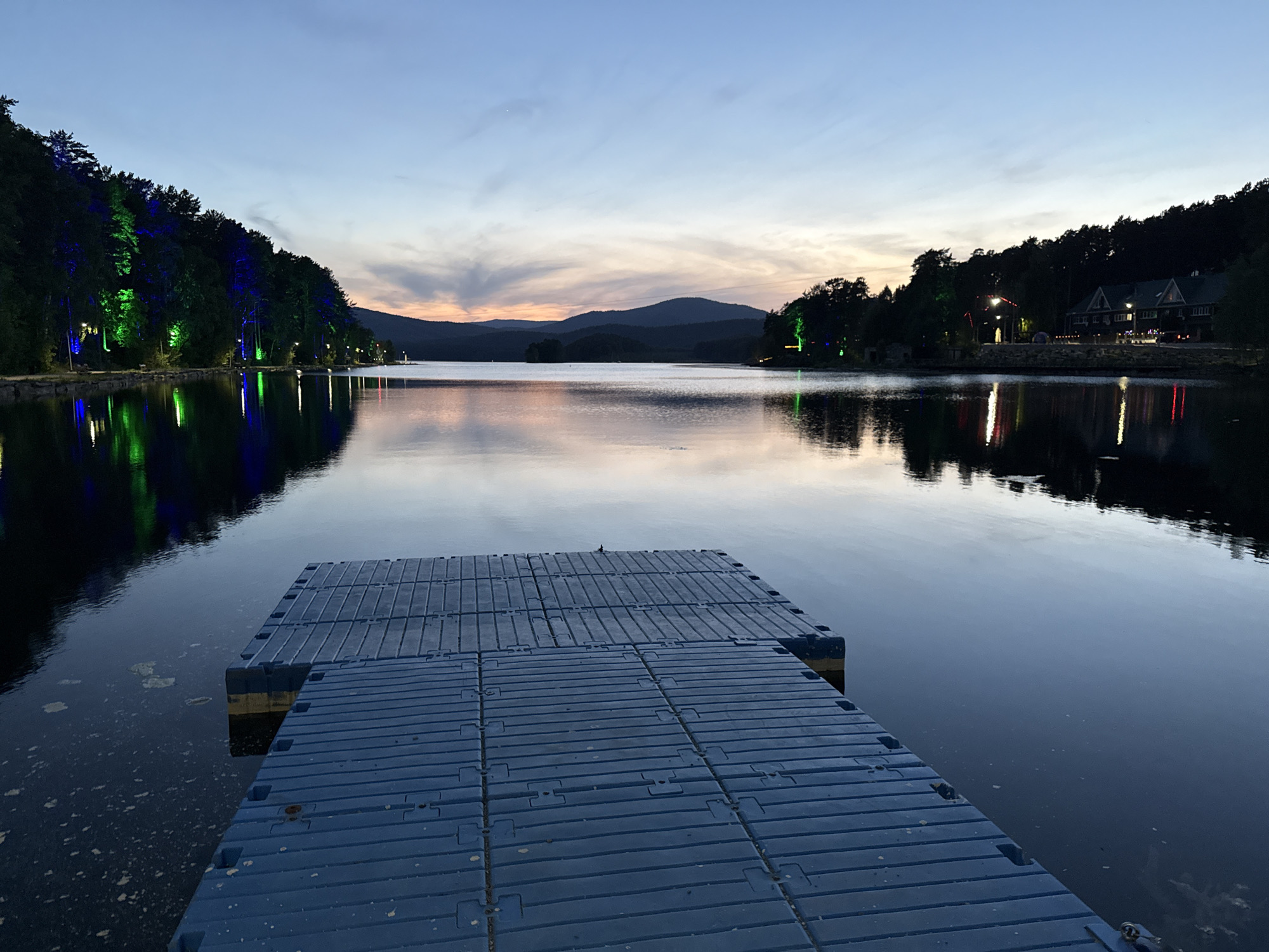 Captured at dusk in Dom Otdykha Zolotoy Plyazh, Russia, the image features a serene lake reflecting the twilight sky and the surrounding landscape. A dark blue floating dock extends from the bottom center of the frame, creating a sense of depth and leading the eye into the scene. On the left and right shores, lush trees line the water's edge. The trees on the left are illuminated with blue and green lights. On the right side, buildings are visible amongst the trees with lights shining. In the distance, silhouettes of mountains can be seen beneath the colorful sky, adding to the picturesque and tranquil atmosphere. The lake's surface is mostly still, enhancing the mirror-like reflection.