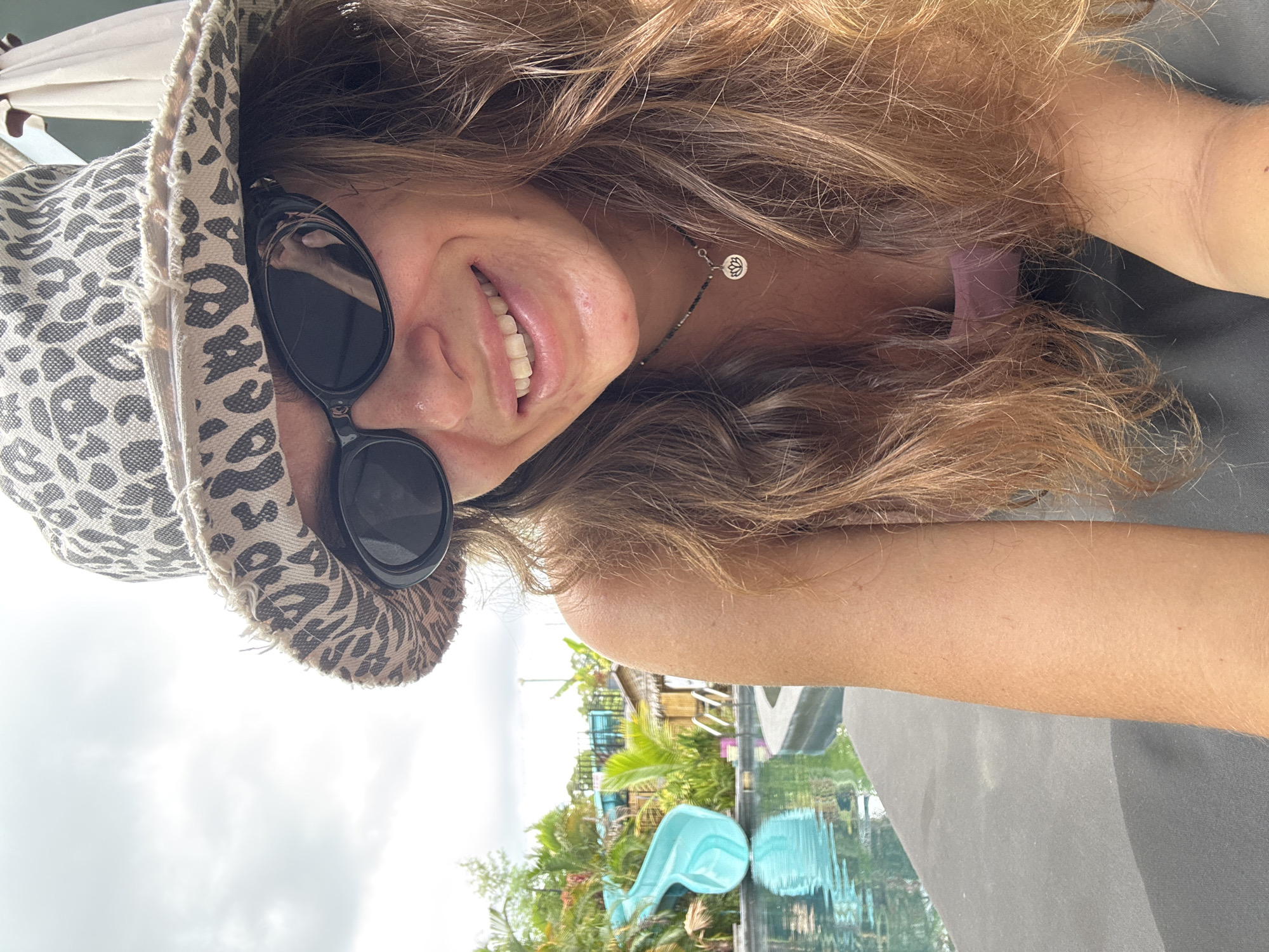 A woman takes a selfie, angled from above, while seated next to a swimming pool. She wears a leopard-print hat, sunglasses, and a necklace with a lotus pendant. The pool reflects nearby buildings and palm trees, placing the location within Baan Mae Nam, Thailand. The sky is overcast.