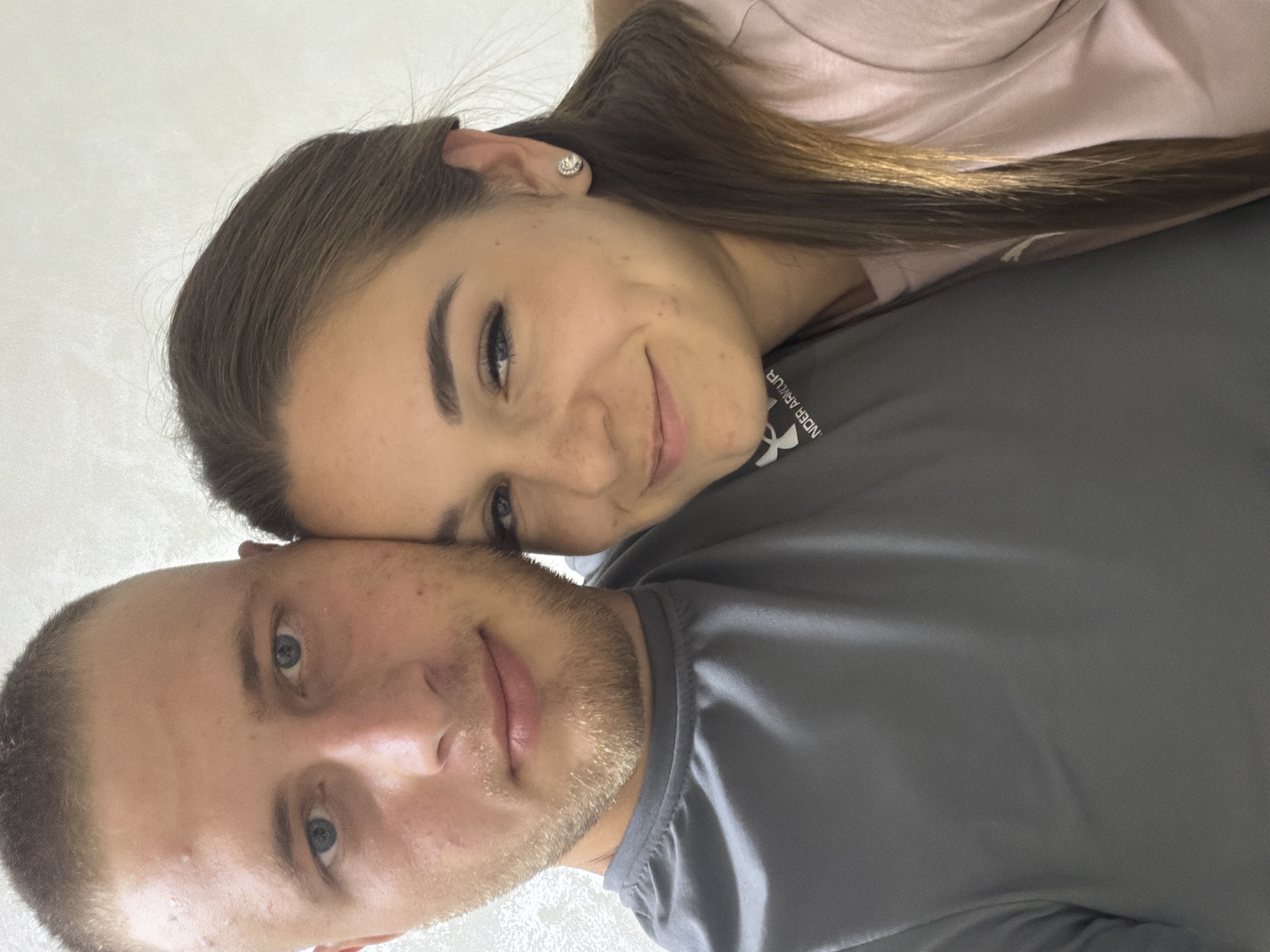 This is a close-up, slightly blurry, horizontally oriented selfie of two people, a man and a woman, with their faces angled towards the camera. The man is in the foreground, his head and shoulders visible. He has light skin, blue eyes, and short, buzzed brown hair. He has a light beard and is wearing a grey t-shirt with the "Under Armour" logo visible on the chest. He appears to be smiling.
The woman is leaning on the man's shoulder and head, her face positioned above and to the side of his. She has fair skin with freckles and blue eyes. Her dark brown hair is long and appears to be styled loosely. She is wearing a light pink shirt and a small, sparkling earring in her visible ear. She is also smiling, with her eyes looking towards the camera.
The background is a plain, light-colored textured surface, possibly a wall. The lighting suggests it's daytime, and there are no obvious clues about the weather or specific location beyond the provided context of Strășeni, Moldova. The overall mood conveyed by the subjects' expressions is affectionate and happy. The image is an informal selfie, likely taken in a casual setting, perhaps at home or outdoors. The visible text on the man's shirt reads "UNDER ARMOUR".