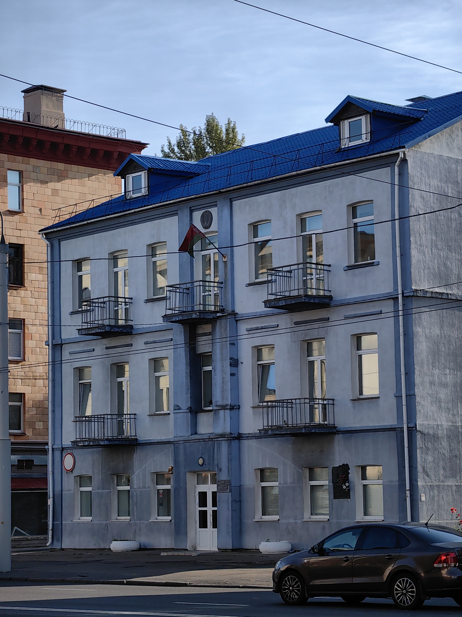 The image shows a four-story building with a blue roof, located in the city of Minsk, Belarus. The building has a light blue facade with several windows and balconies on each floor. A flag, appearing to be the flag of Belarus, is displayed on the right side of the building. To the left of the building, a taller brick building is visible. A dark-colored car is parked in front of the blue-roofed building. The scene appears to be a street in a city, with power lines and a traffic light visible. The sky is clear and blue.